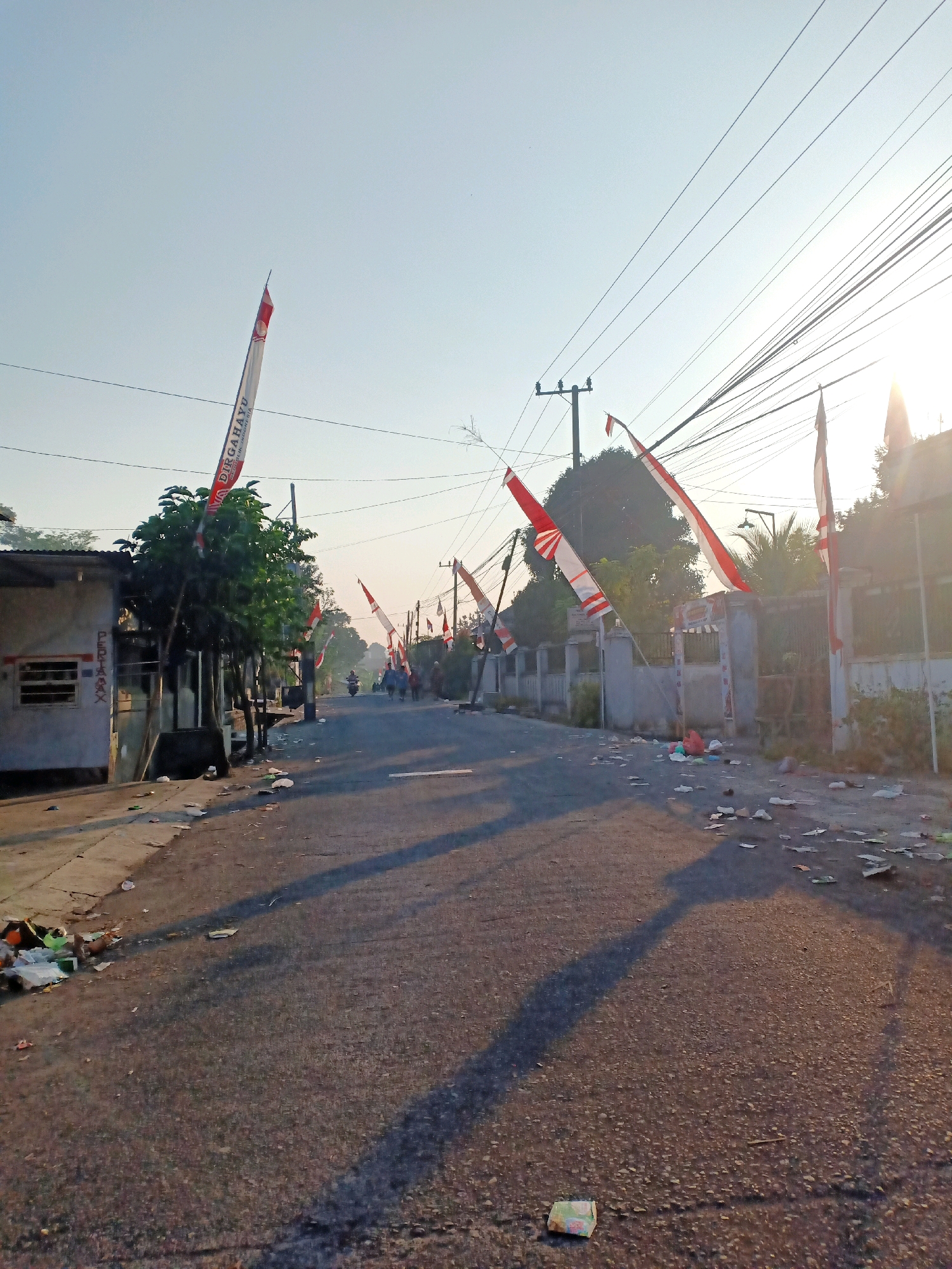 The image shows a street in Kalibendo, Indonesia, decorated with Indonesian flags for a national celebration. The flags, in red and white, are hung on poles and wires along the street. The scene is bathed in the warm light of what appears to be sunrise or sunset, casting long shadows across the asphalt road. The street is littered with various pieces of trash, including plastic bags and paper, suggesting recent activity or a lack of regular cleaning.
On the left side of the street, there is a low-rise building with a corrugated metal roof and a window covered with bars. Adjacent to it, a leafy tree stands tall, its branches partially obscuring the flags above. Further down the street, several people are visible, some on motorcycles and others walking, moving away from the camera.
The environment appears to be a residential or semi-commercial area, with houses and possibly small businesses lining the road. Power lines crisscross the sky, supported by wooden poles. The overall impression is of a community preparing for or celebrating a national event, despite the presence of litter.