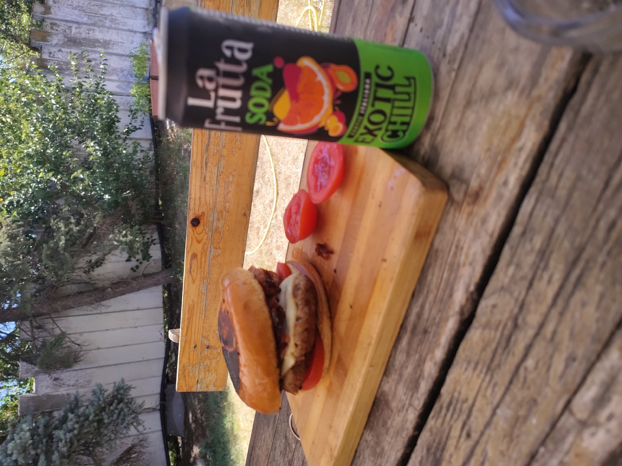 A close-up, eye-level shot shows a burger and a can of "La Frutta SODA Exotic Chill" on a wooden picnic table. The burger is placed on a wooden cutting board, with two tomato halves to its right. The can of soda is black with a green base, featuring colorful fruit illustrations and text. In the background, a wooden fence and lush green trees are visible under a bright sky. The scene appears to be outdoors, possibly in a backyard or park, with a relaxed, casual atmosphere.