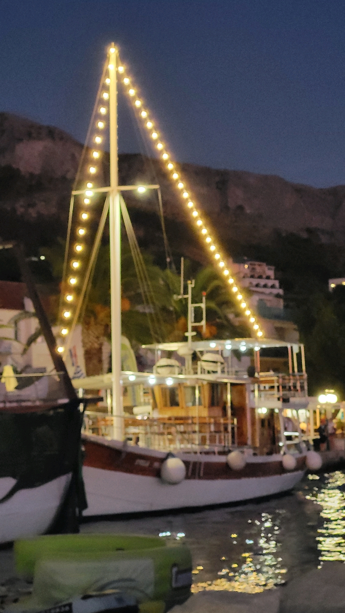 A nighttime view of a boat adorned with string lights is docked in a harbor, likely in Općina Tučepi, Croatia. The boat, with a prominent mast and a white hull trimmed with red, has multiple decks and windows. The string lights ascend the mast in a triangular pattern, illuminating the scene against a dark sky and a silhouetted hillside with buildings. The water reflects the lights, creating a shimmering effect. In the foreground, another boat or marine equipment is partially visible, rendered in a blurred, lime-green hue. The overall atmosphere is tranquil, with a hint of festivity due to the decorative lights.