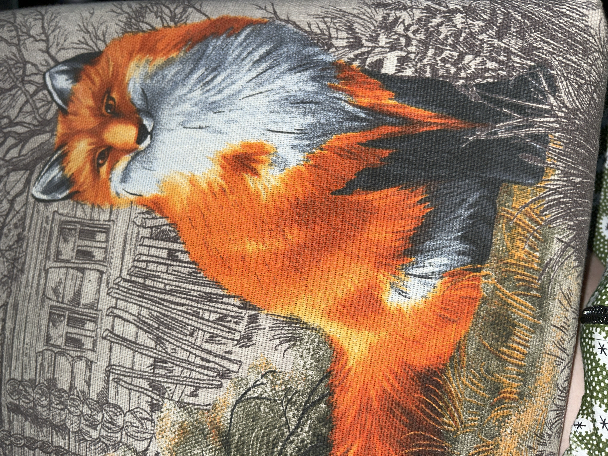The image displays a close-up of a fabric with a printed design of a fox in a naturalistic setting. The fox is depicted in vibrant orange, white, and gray colors, with its head turned towards the viewer. Behind the fox, the design includes what appears to be a rustic wooden structure with windows, possibly a house or cabin, and barren trees. The ground is rendered with textured grass and foliage. The fabric itself has a visible woven texture. There are no people or discernible actions depicted in the image. The location within the city of Tugulym, Russia, is not inferable from the provided image.