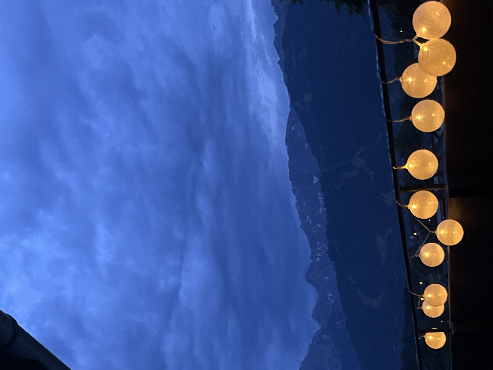 The image depicts a twilight scene with a string of illuminated, globe-shaped lights suspended over a railing. In the background, dark, layered mountain ranges are visible against a cloudy, deep blue sky. The setting suggests an outdoor balcony or patio with a view of the mountains. The overall atmosphere is tranquil and serene, characteristic of an evening in a mountainous region such as Garmisch-Partenkirchen, Germany. No people or specific actions are captured in the frame.