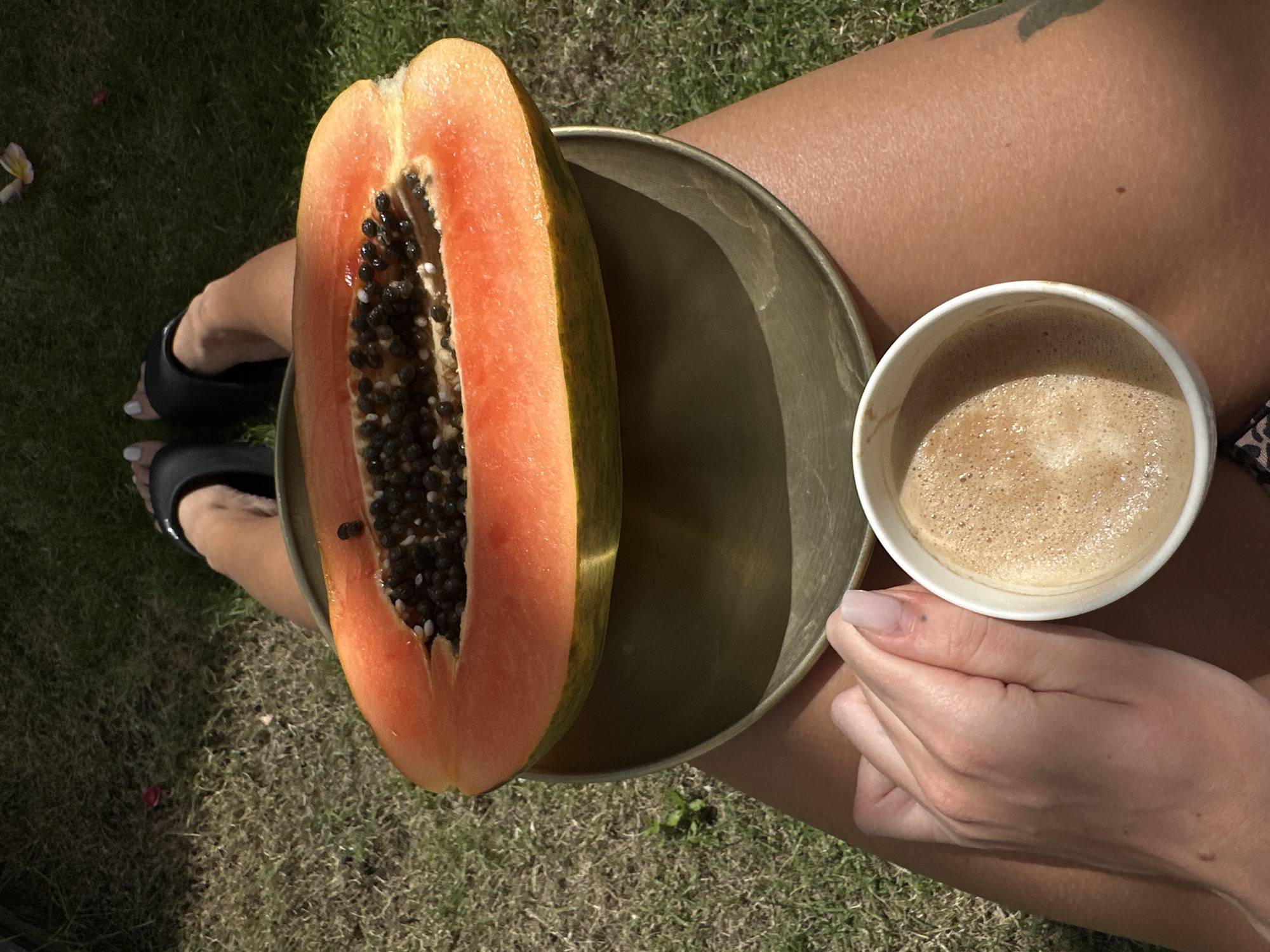 A top-down shot captures a woman seated on grass in North Kuta, Indonesia. On her lap rests a round, shallow, possibly bronze-colored plate holding a halved papaya with black seeds. Adjacent to the plate is a white cup filled with coffee. Her feet are visible, shod in black sandals. Her hands, one holding the cup and another resting next to it, are pale and manicured. The background consists of green grass and partial shade.