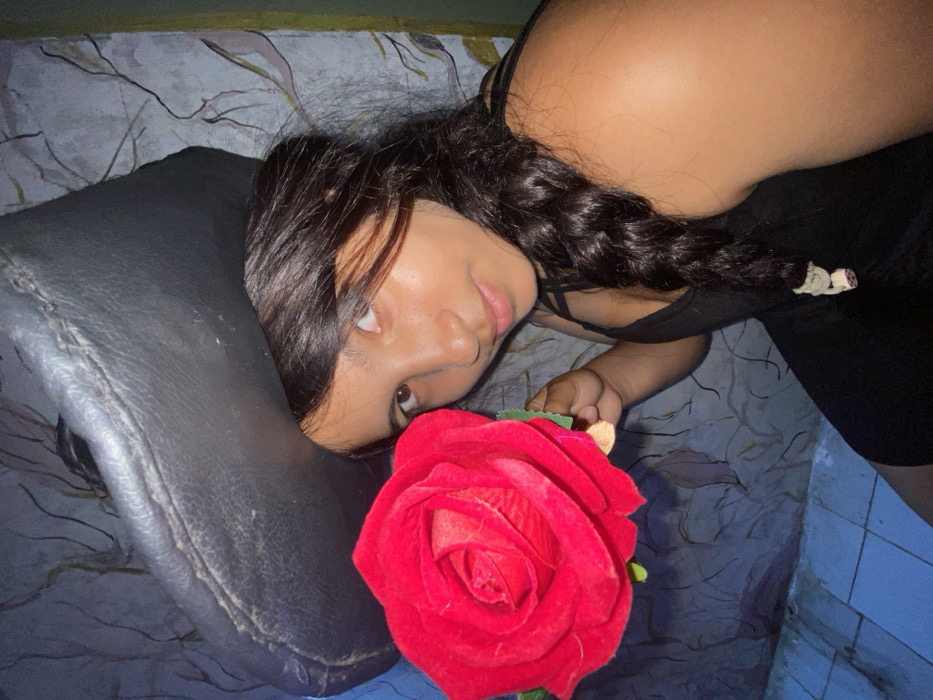 A woman lies on a patterned surface, resting her head on a black cushion. She holds a large red rose with green stem in her hand, positioned near her face. She has dark hair, styled with loose braids, and wears a black top. A glimpse of blue tiled wall is visible in the corner, suggesting an indoor setting.