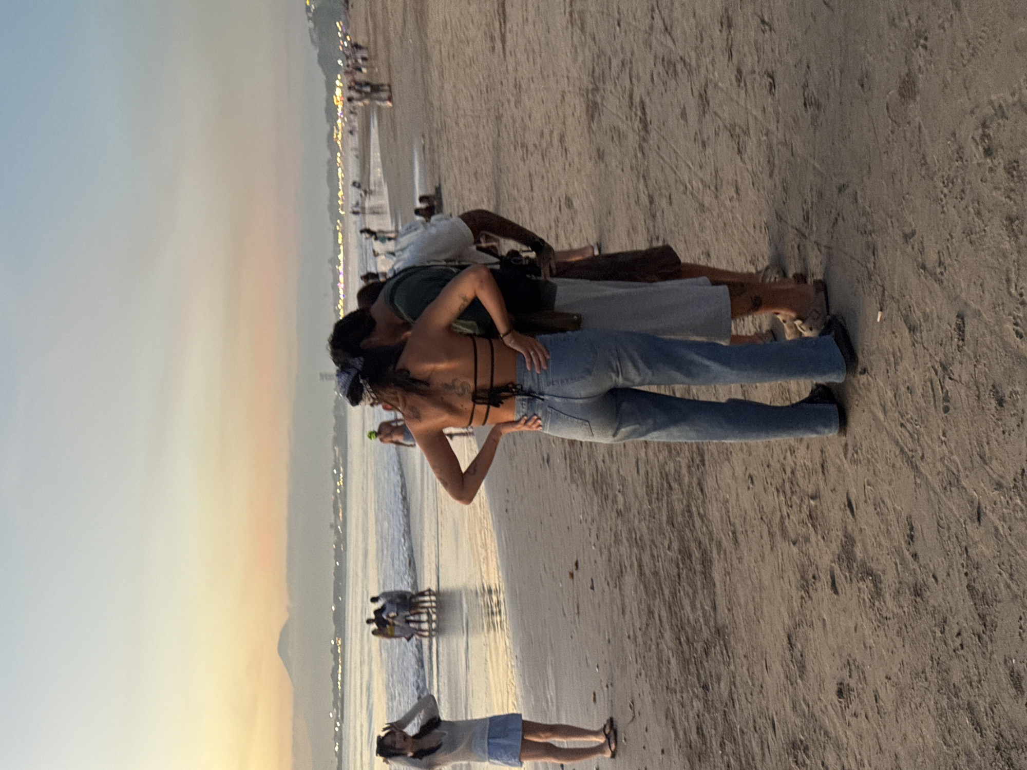 The image captures a beach scene in Kuta Selatan, Indonesia at dusk. The sky is tinged with hues of orange and yellow. The sand is light brown and appears slightly damp near the water. Several people are present on the beach. In the foreground, a woman wearing jeans and a top with straps is standing with her back to the camera, interacting with others. Nearby, another person lies on the beach. Further towards the water, there are more people gathered, some appearing to be sitting in chairs. The water is calm, and in the distance, there is a faint outline of a hill. Along the shoreline, there is a line of lights.