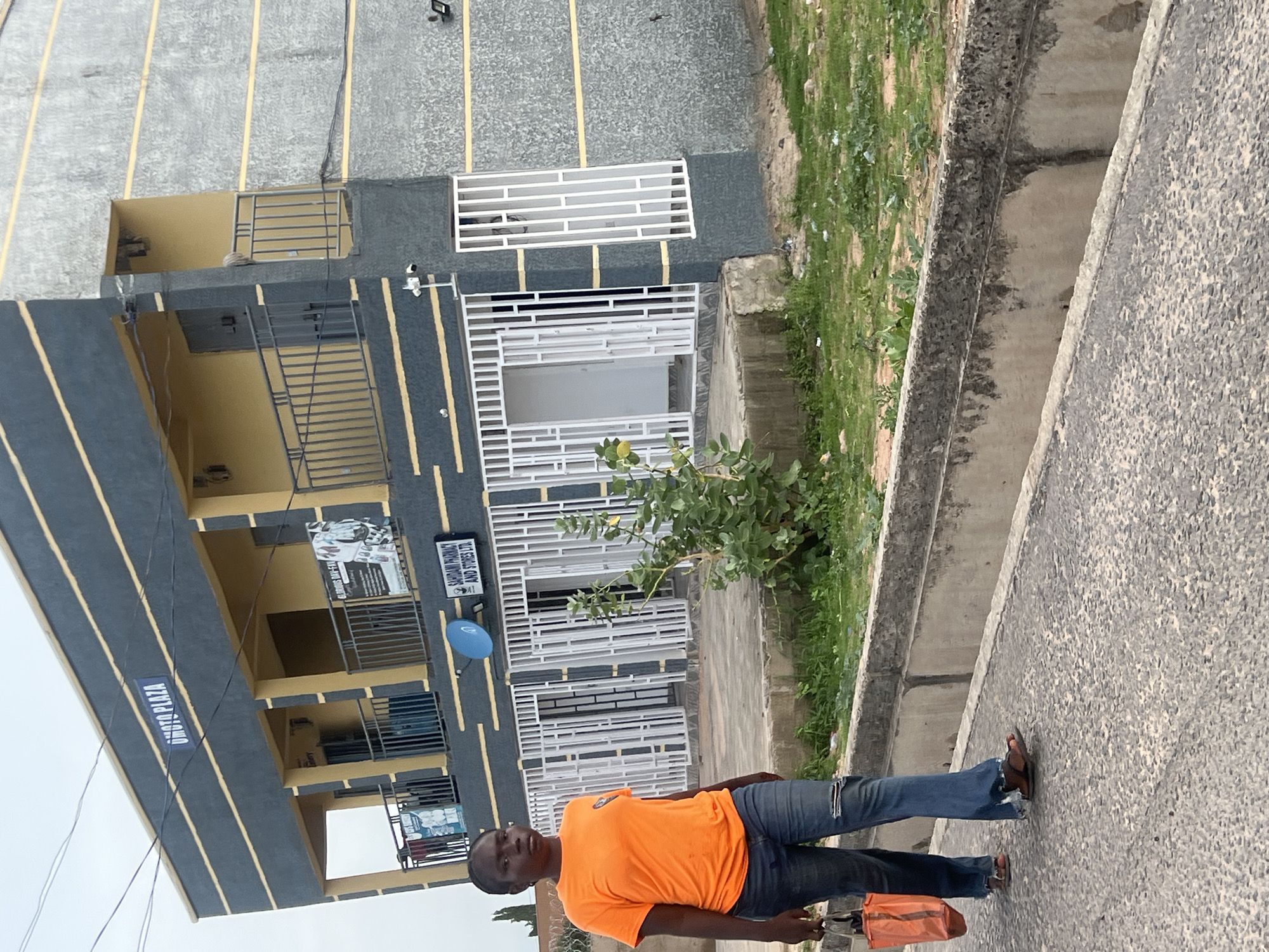 The image, taken in Howayi, Nigeria, captures an outdoor scene featuring a multi-story building and a person standing on a paved surface. The building, on the left, has a grey and dark grey facade with distinctive yellow horizontal stripes. It features numerous windows and balconies protected by white and dark metal grilles. A blue satellite dish is mounted on the building's exterior.
A man, wearing an orange t-shirt, blue ripped jeans, and flip-flops, stands in the foreground, holding a small orange bag. He looks towards the camera with a neutral expression. To the right of the paved area, a concrete curb separates it from a grassy patch with sparse vegetation. The sky appears overcast, indicating daytime.
Visible text on the building includes a prominent sign reading "OMOTO PLAZA". Below it, another sign states "SAHDAN PHARMACY AND STORES LTD".