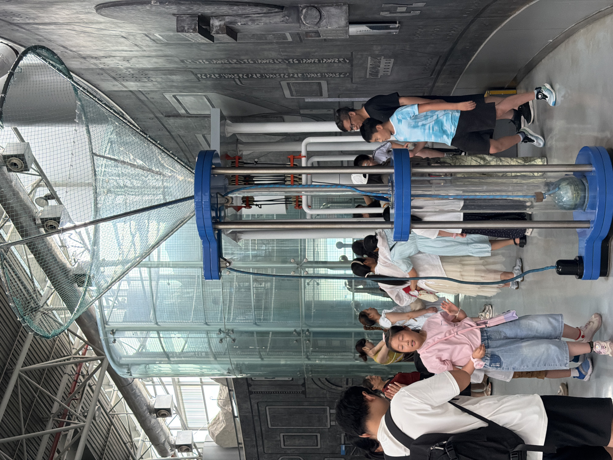 In an indoor exhibition space within Jiangbeicheng Subdistrict, China, a group of people, including adults and children, are gathered around a large, blue, metal and plastic exhibit. The exhibit features a glass tube in the center, connected to tubes and pulleys at the top and bottom. A green net structure extends upwards from the top of the exhibit. To the left of the exhibit is a tall, cylindrical glass structure, possibly part of another exhibit or architectural element, behind which is a large window showing a bright, overcast sky. The background consists of a dark gray, textured wall with various rectangular and circular indentations, resembling panels. The floor is a smooth, light gray material. Several individuals are looking at the exhibit or interacting with each other. A young girl in a pink shirt and denim shorts is in the foreground, looking towards the exhibit and gesturing with her hands. A young boy in a blue tie-dye shirt and black shorts is standing to the right of the exhibit, also looking at it.