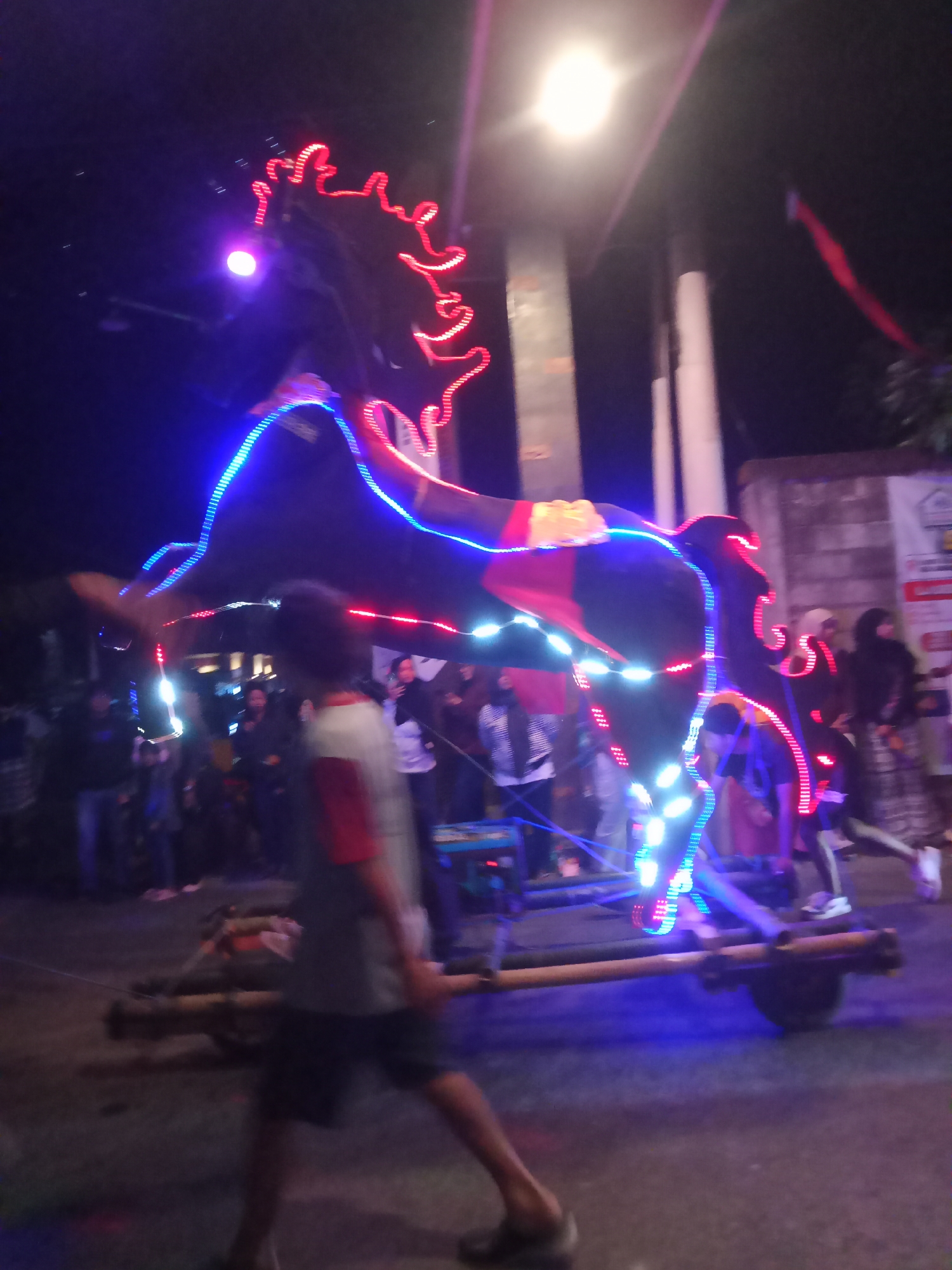 In Urutsewu, Indonesia, at night, a large, dark horse effigy is illuminated with red and blue LED lights. The effigy is mounted on a wooden cart with two wheels. A person wearing a grey t-shirt with red sleeves and black shorts walks in the foreground, partially obscuring the cart. In the background, a crowd of people are visible, as well as buildings and streetlights. The scene appears to be a night-time event or festival.