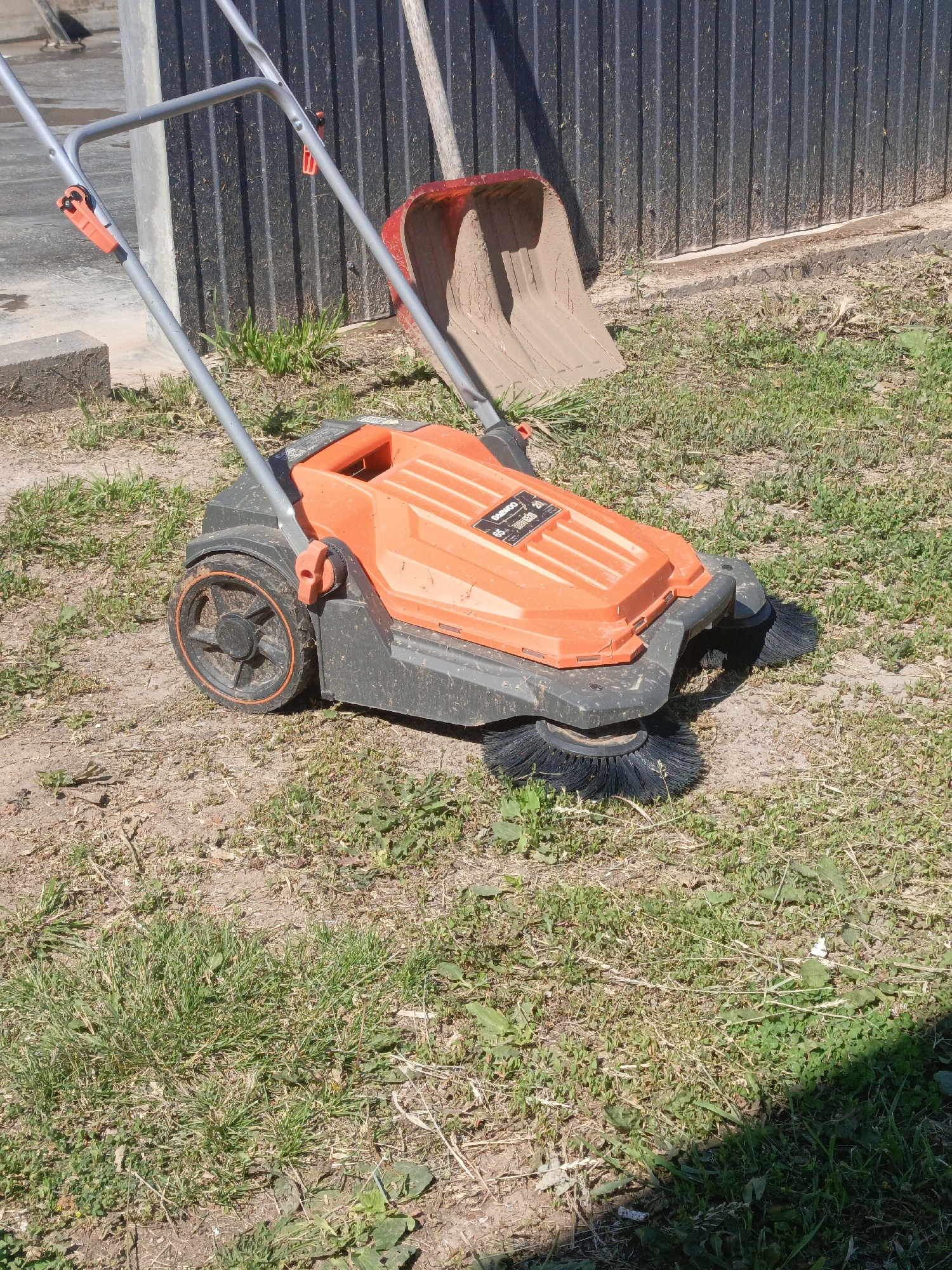 An orange and black mechanical sweeper sits on dry, patchy grass and dirt. The sweeper has two rotating brushes at the front and a handle with orange grips. Behind the sweeper, leaning against a dark corrugated metal fence, is a red shovel. The scene is outdoors, with a concrete structure visible to the left. The ground is mostly dry earth with sparse grass and weeds. The lighting suggests a sunny day.