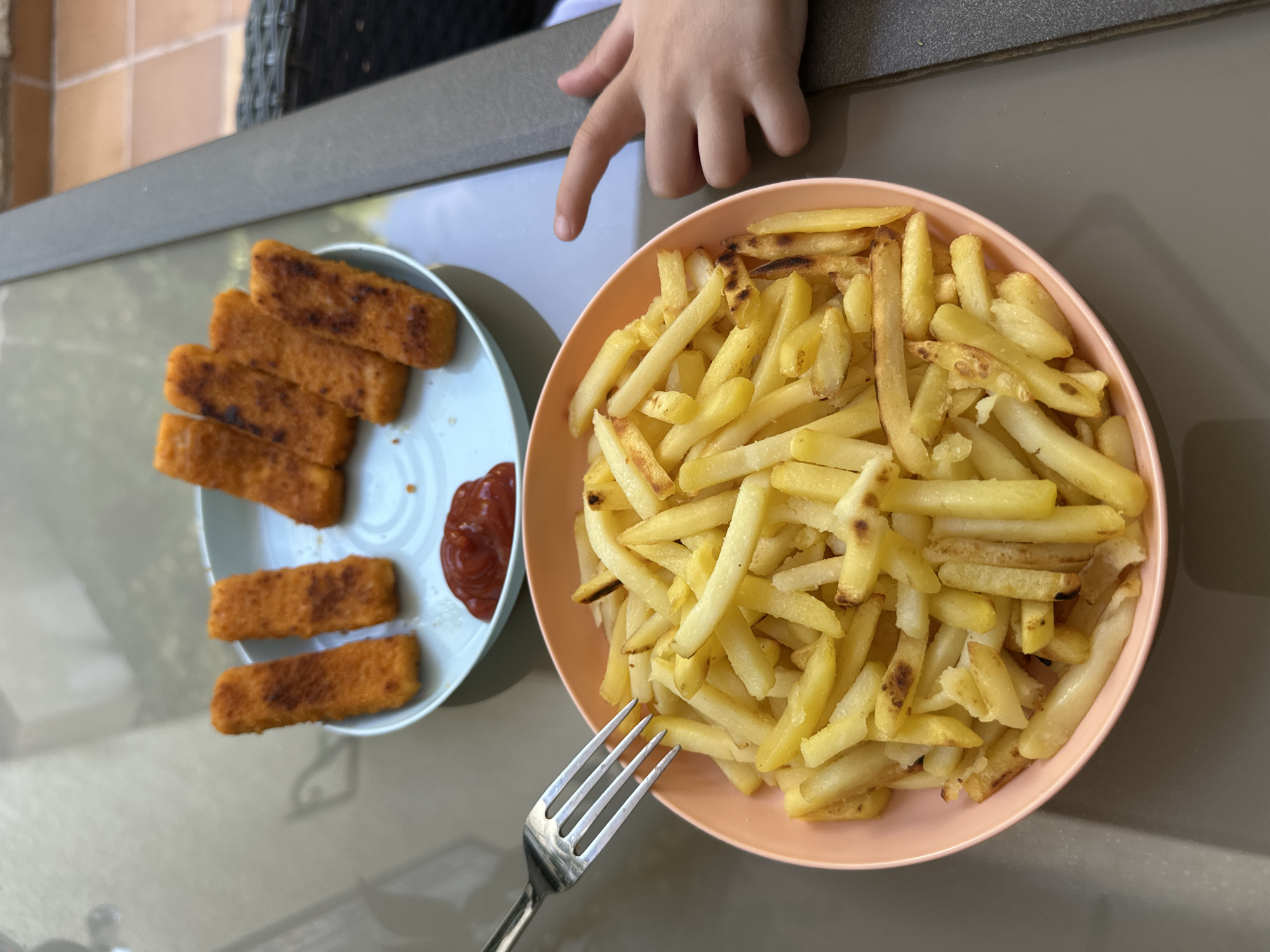 A child's hand is pointing towards a plate of fish fingers and a bowl of french fries on a glass table. The fish fingers are golden brown and arranged on a light blue plate with a dollop of ketchup. The french fries are golden yellow and piled high in a peach-colored bowl. A fork rests on the edge of the bowl, with some fries on it. The scene appears to be outdoors, possibly on a patio, with a glimpse of tiled flooring in the background. The child's hand is positioned to the right of the frame, with the index finger extended. The overall impression is a casual mealtime setting. The location is specified as Sant Feliu de Guíxols, Spain.