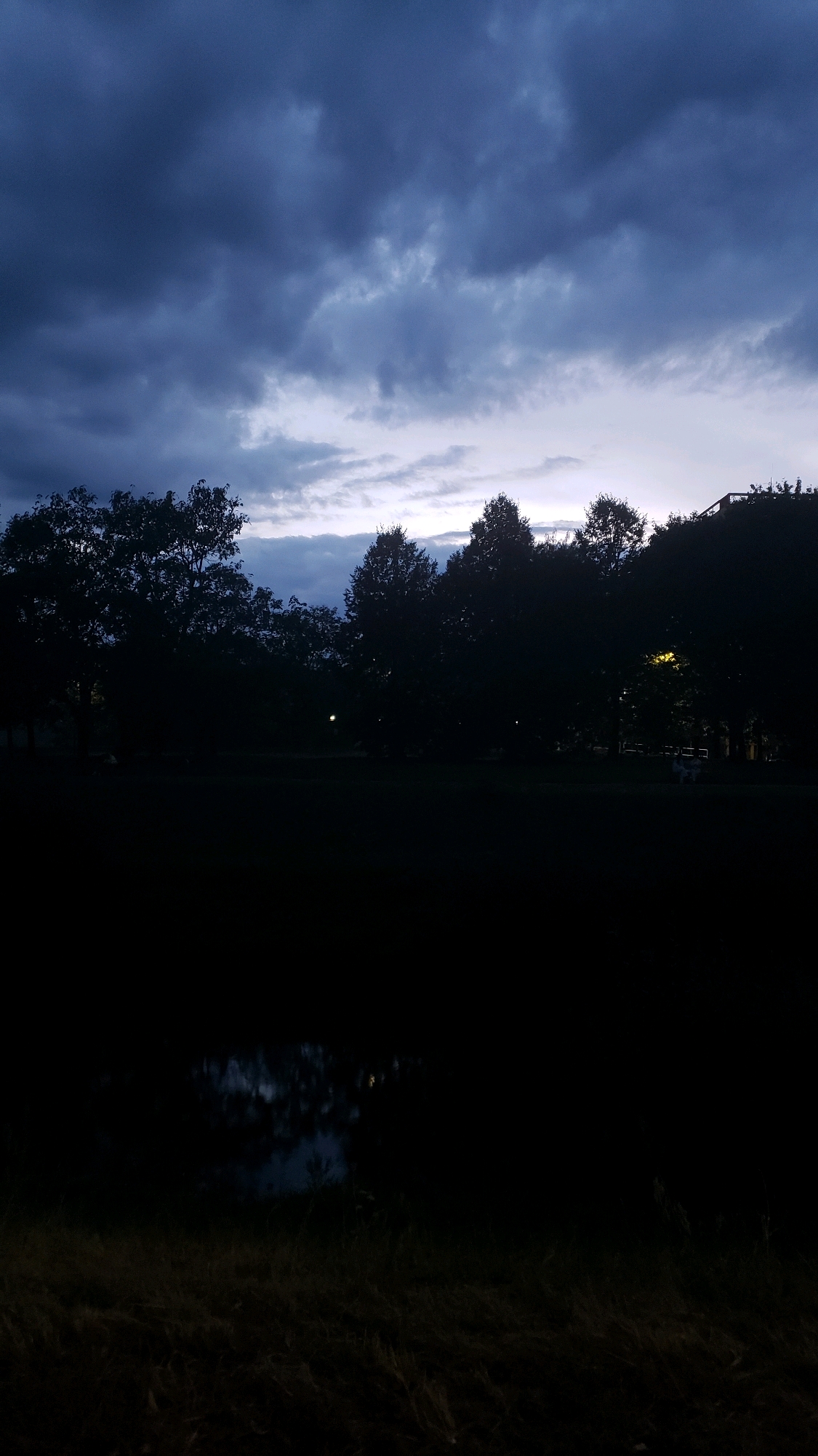 A wide-angle, full shot captures a dusky scene with dark, silhouetted trees against a stormy, twilight sky. The sky is filled with heavy, dark blue clouds, with streaks of lighter grey and pale blue peeking through, suggesting the last remnants of daylight or the onset of night. A dark, still body of water reflects the sky's muted colors in the foreground. The middle ground is dominated by a dense line of trees, their foliage rendered as solid black shapes due to the low light conditions. Faint lights from what appear to be streetlights or distant buildings are visible through the trees, casting a subtle glow. The ground in the very foreground is covered in dry grass and appears dark and indistinct. The overall atmosphere is somber and perhaps indicative of impending rain or the end of a cloudy day. The location is identified as the park area near the main road to Prešov, Slovakia, specifically near the area of the former military barracks.