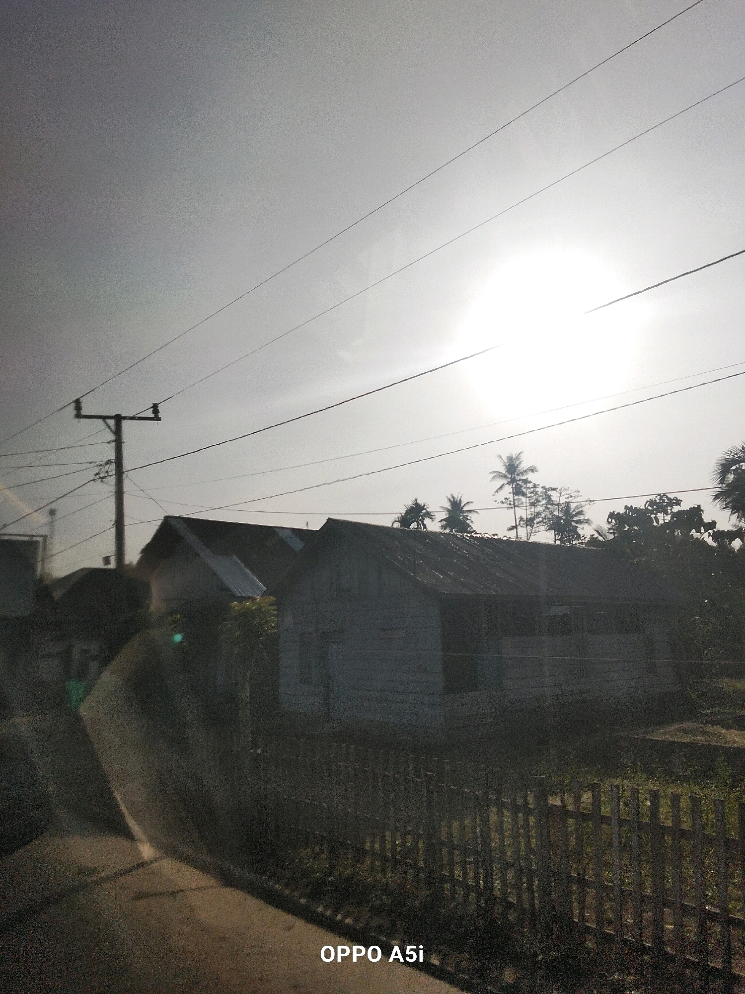 In Tabanggele, Indonesia, an overcast sky dominates the upper half of the image, with a bright, diffused sun positioned slightly to the right. Several power lines crisscross the sky, appearing as thin, dark lines against the bright background. In the middle ground, a single utility pole with an attached crossarm stands prominently. To the right of the pole, two low-slung, elongated houses are visible. The house in the foreground has a corrugated metal roof and a wooden fence in front of it. The house behind it is partially obscured. Trees, including palm trees, are sparsely scattered behind the houses. The lower portion of the image shows a dark, paved surface, possibly a road or pathway, leading towards the foreground. The overall lighting suggests it is either early morning or late afternoon. The "OPPO A5i" watermark is visible in the lower right corner.