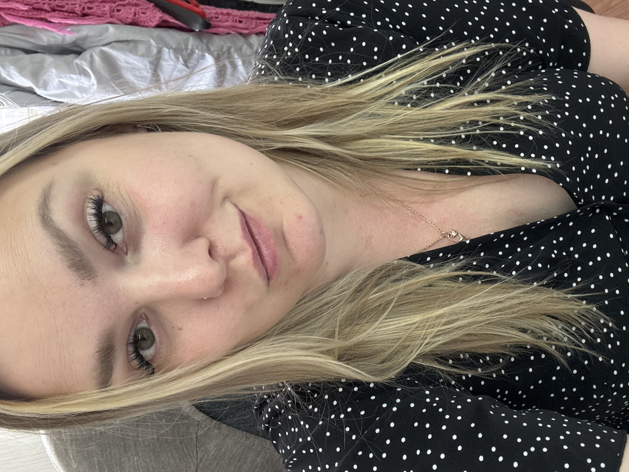 A woman with blonde hair and light skin is lying on a bed, wearing a black dress with white polka dots. A necklace is visible around her neck. The bedding consists of a gray sheet and a pink knitted blanket. There is no evidence to suggest this is in Noviy, Russia.