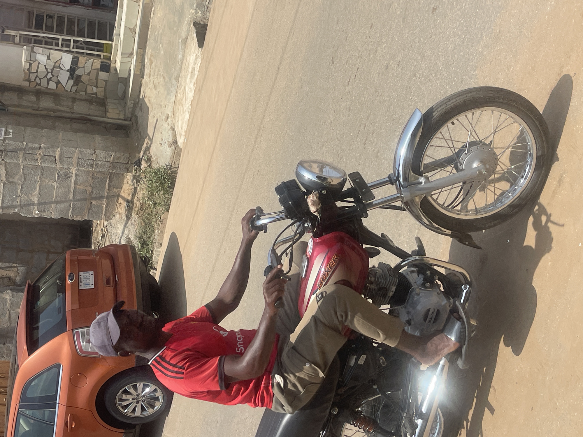 A man wearing a red t-shirt and a grey baseball cap rides a red motorcycle down a paved road. The man is wearing khaki pants and appears to be wearing sandals. He is looking forward, with his hands on the handlebars. The motorcycle is a Boxer brand. In the background, an orange SUV is parked on the side of the road. The environment appears to be urban, with buildings made of brick and stone. The sky is bright and sunny, suggesting it is daytime. There are some sparse green plants growing on the side of the road. This scene is likely taking place in Abuja, Nigeria, given the visual cues often associated with urban environments in that region, such as the architecture and vehicle types.