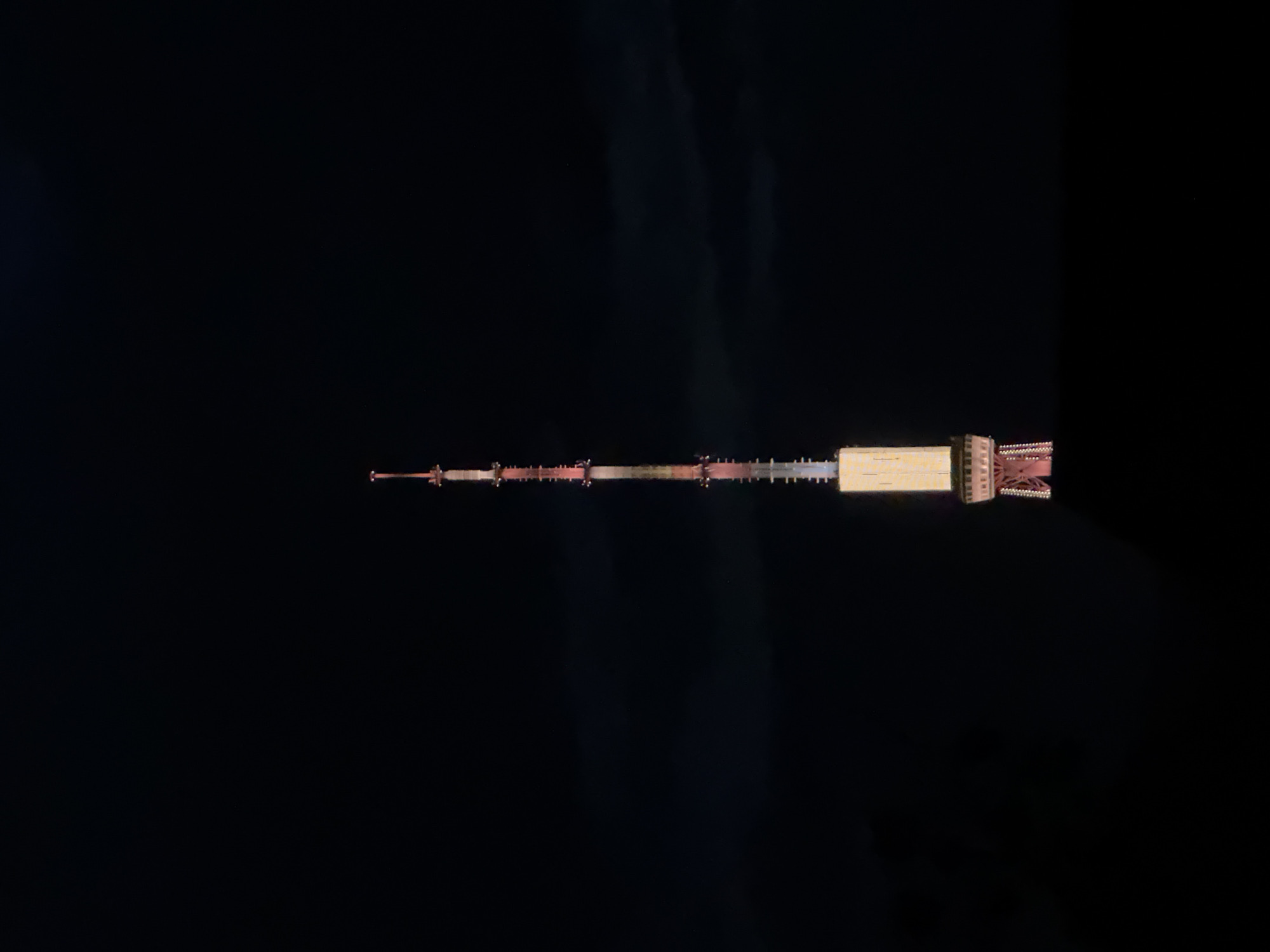The image depicts the Yerevan TV Tower at night, viewed from above, possibly from a drone. The tower is brightly lit, standing out against the dark sky. It features a tall, slender structure with multiple levels and architectural details at the base. The surrounding area is obscured by darkness, but there are some hints of clouds. The tower is located in Yerevan, Armenia.