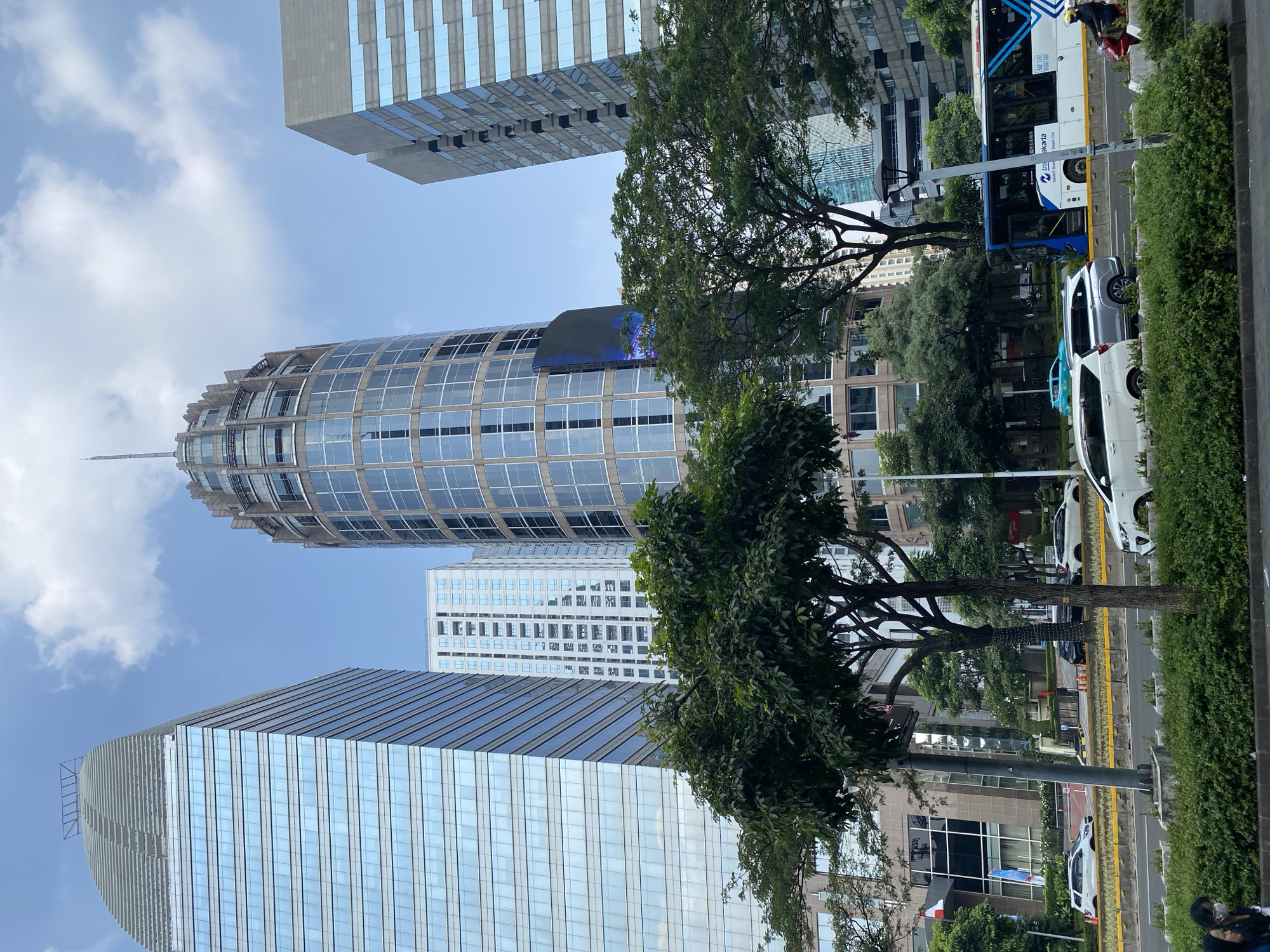 In Central Jakarta, Indonesia, a busy street scene unfolds with towering modern buildings dominating the skyline. On the left, a glass-clad skyscraper reflects the blue sky and clouds. To its right, a distinct cylindrical building with a decorative crown and a pointed spire rises prominently. Further right, another rectangular skyscraper with blue-tinted windows stands tall. In the foreground, a paved road is lined with lush green trees and bushes. Vehicles, including a white SUV, a silver SUV, and a white sedan, are on the road. A blue and white bus with "Jakarta Smart City" emblazoned on its side is also visible. A person on a red motorcycle, wearing a yellow helmet, is seen in the distance. Two pedestrians are in the lower left corner, one wearing a face mask. The sky is partly cloudy with patches of blue.