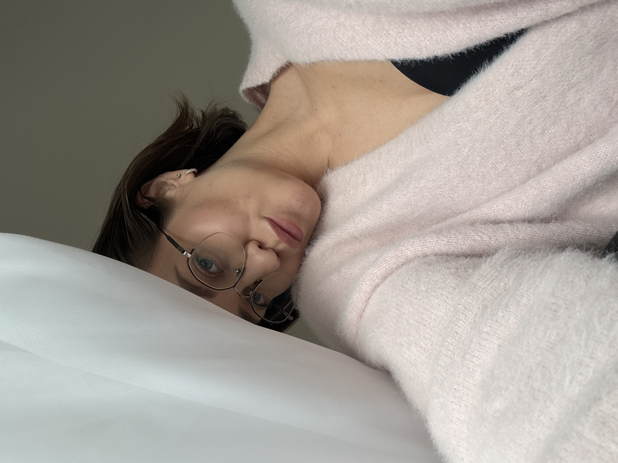 A woman wearing glasses and a pink sweater is lying on a white surface, possibly a bed or a similar soft, flat area. The angle of the photo is taken from above, looking down at the woman. No specific details place the image in Pervomaysky, Russia; therefore, the location cannot be confirmed from the visual data.