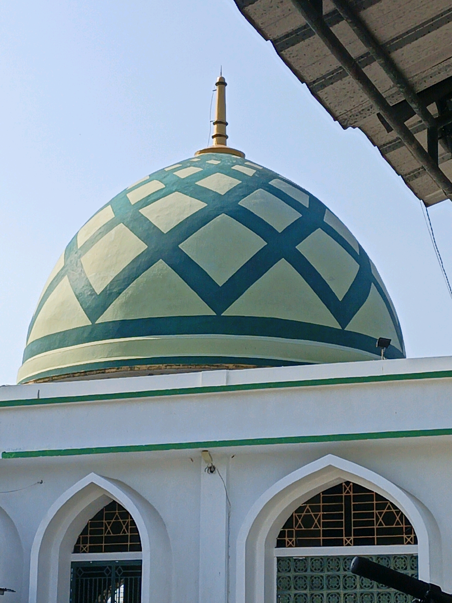 A mosque dome in Kalipurwo, Indonesia, is the subject of this image. The dome is adorned with a grid pattern of dark green against a pale green background, and topped with a gold finial. Below the dome, a white building with arched windows and green trim is visible. The windows are covered with patterned metal grilles. A portion of a dark, corrugated roof structure is visible in the upper right corner of the frame, partially obscuring the sky. The scene is set against a clear blue sky.