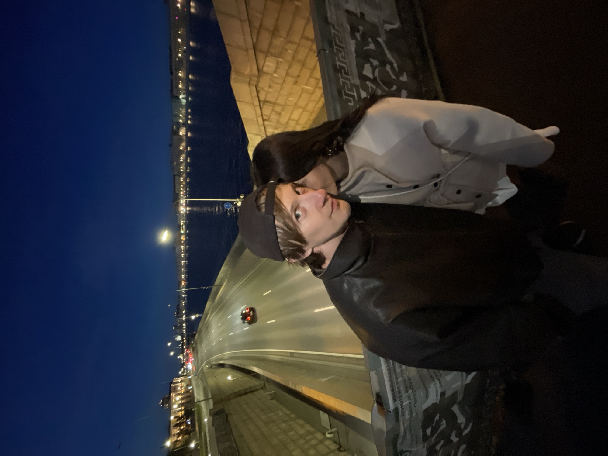This is a high-angle, waist-up, medium shot of a couple on a bridge at night in Saint Petersburg, Russia. The man, wearing a black leather jacket and a baseball cap, faces the camera with a light-colored wall to his right and a dark blue body of water beyond. A woman, facing away from the camera and towards the man, is whispering in his ear. The background features a city skyline with illuminated buildings and streetlights reflecting on the water. A road with a single car driving on it runs parallel to the water on the left side of the image. The bridge railing in the foreground has decorative carvings and text. The lighting suggests dusk or nighttime.