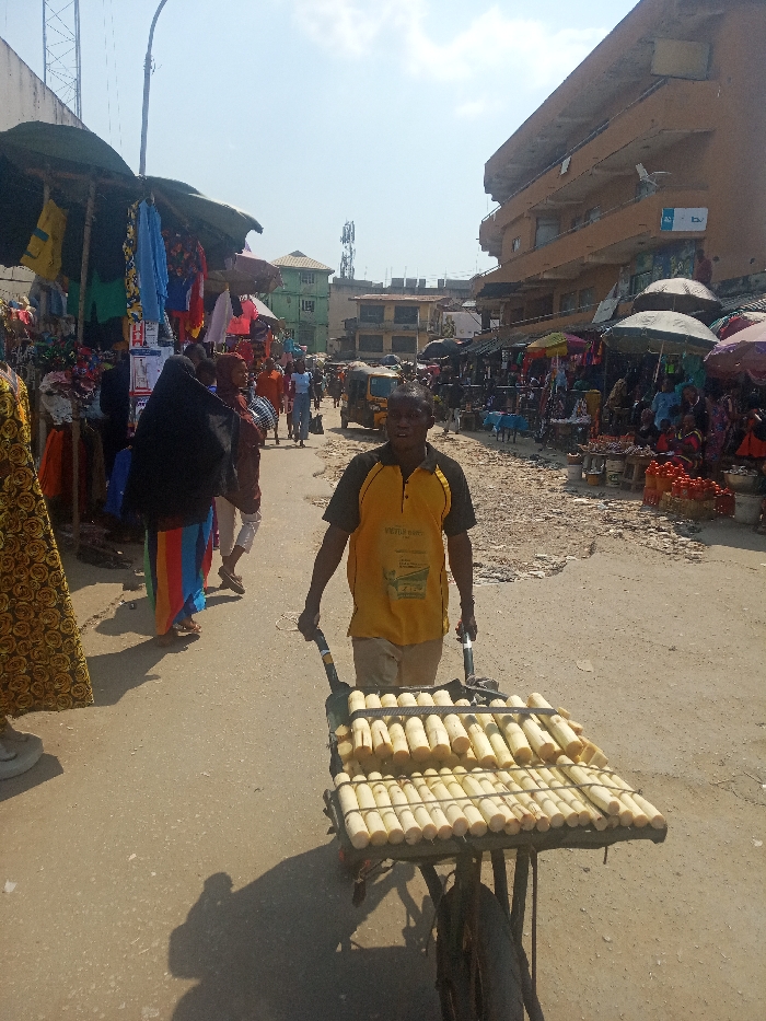 The image displays a bustling outdoor market street in Nkwelle Ezunaka, Nigeria, under direct sunlight. A dark-skinned male, wearing a yellow polo shirt with black accents and light-colored trousers, occupies the foreground, pushing a two-wheeled handcart laden with horizontal rows of peeled sugarcane segments secured by straps. He faces the camera directly. To his left, a market stall under a dark awning displays textiles, with a person in a black hijab and a striped skirt walking away. The street is an unpaved, dusty thoroughfare. To the right, multiple market stalls operate under large umbrellas, featuring goods such as red produce in baskets. In the midground, pedestrians move along the street, and a yellow three-wheeled vehicle is visible. The background is composed of multi-story buildings, including a prominent light-brown structure on the right and a lighter building to the left, adjacent to a communication tower. Sharp shadows indicate clear, sunny conditions.