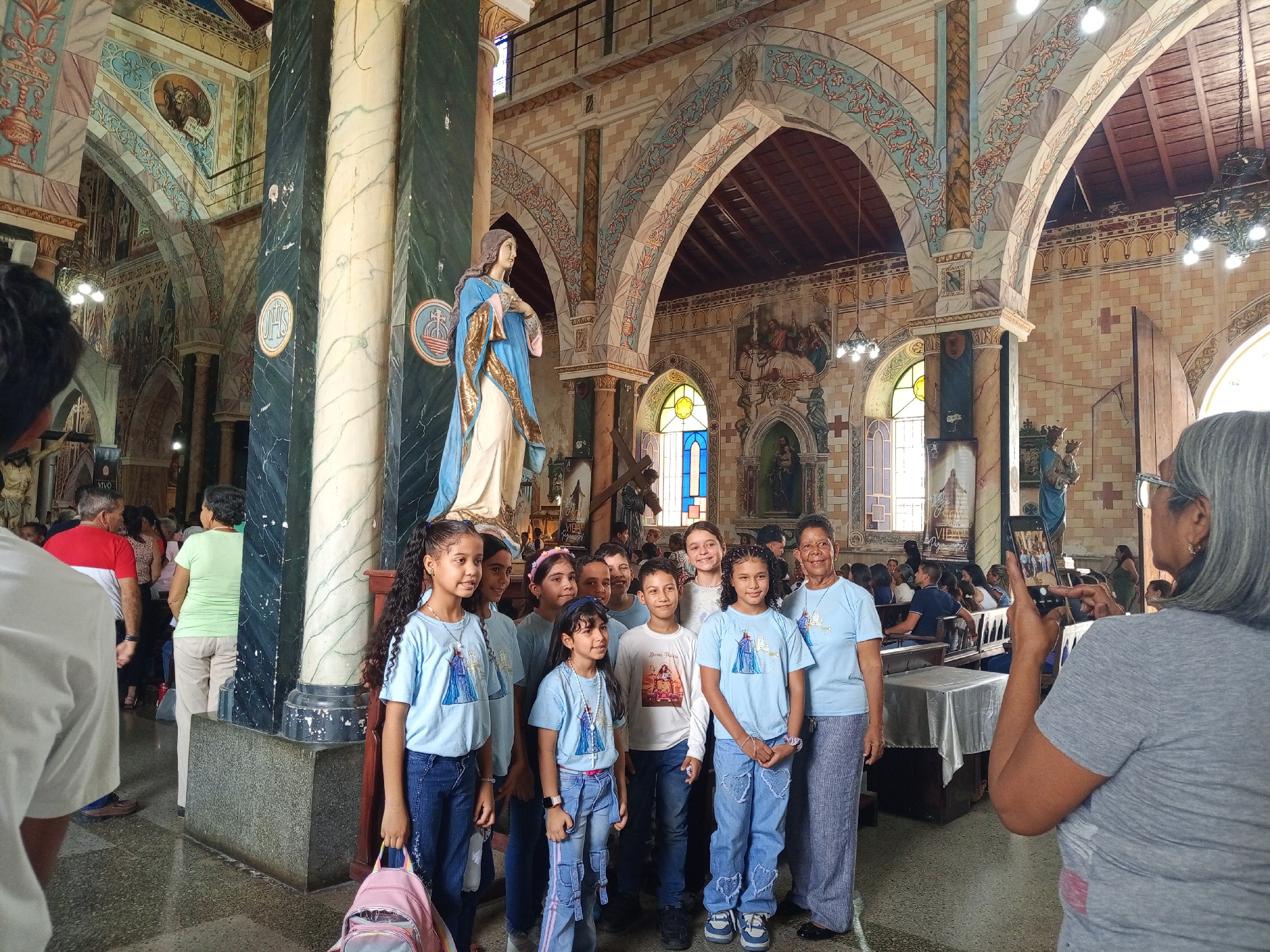 A group of children and an adult are being photographed inside the Iglesia Inmaculada Concepción in Tinaquillo, Cojedes, Venezuela. The group of children and one adult are standing in front of a statue of a woman, which is on a pedestal next to a marble column. The children are wearing t-shirts, some with a graphic of a woman in a dress, and jeans. The adult is wearing a similar t-shirt and pants. A woman is taking a photo of the group with a smartphone. Other people are visible in the background, including other attendees of the church. The church has arches and painted walls.