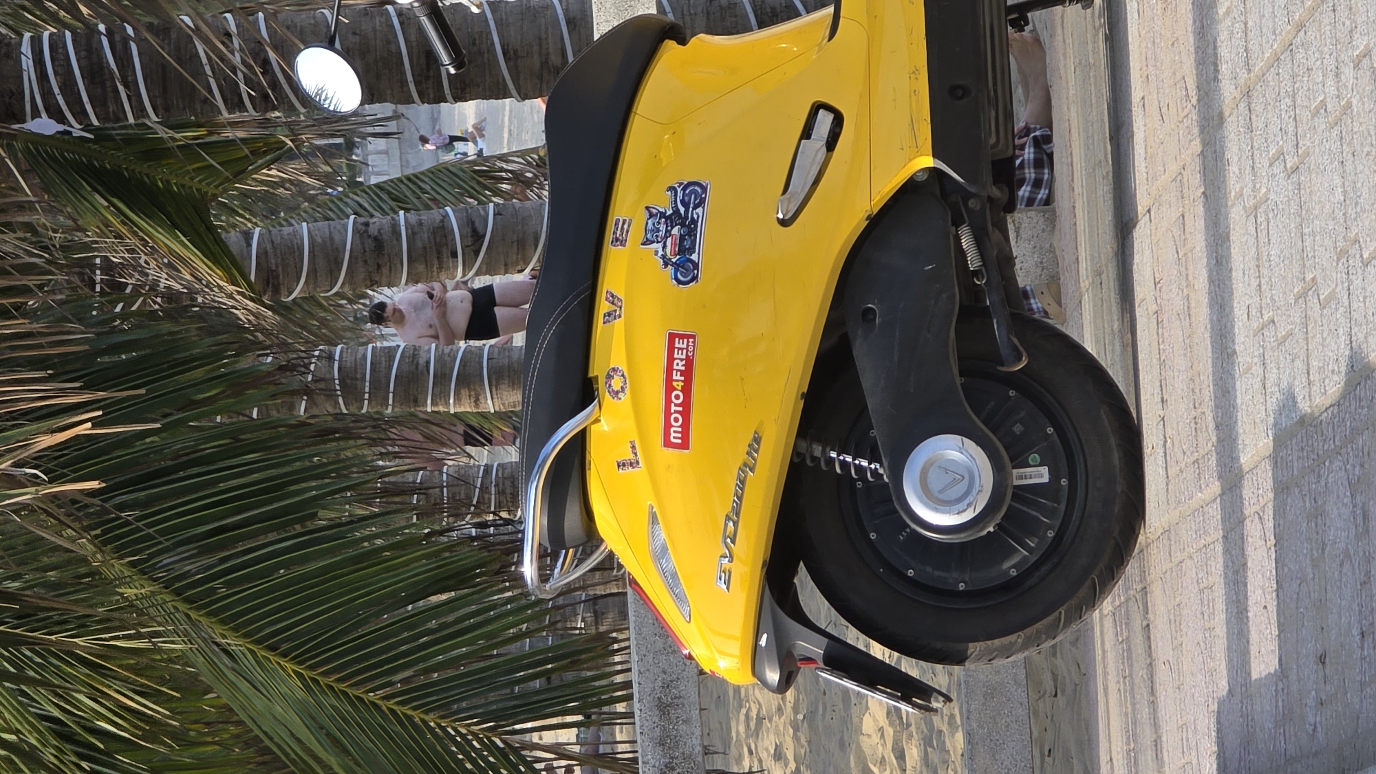 The scene depicts a yellow EV Landlite scooter parked on a stone walkway in Nha Trang, Vietnam. The scooter is parked near palm trees, and in the background, there is a person sunbathing on the beach. The environment appears to be a typical beach scene.
