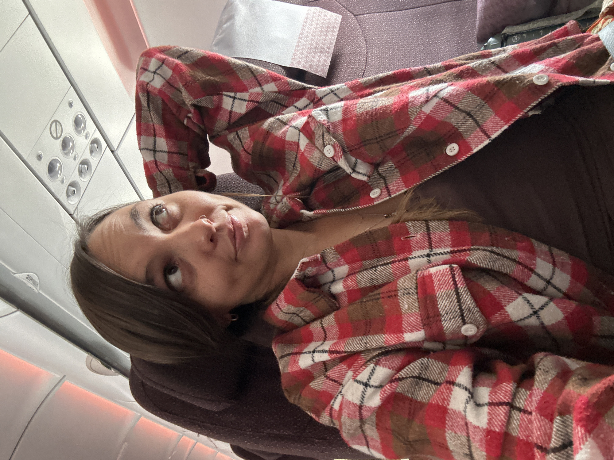 The image shows a woman lying in an airplane seat. She has brown hair and is wearing a brown shirt and a red and white plaid flannel shirt. Her head is tilted back, and she is looking up and to the left. The airplane window and overhead controls are visible in the upper left corner of the image. The background is a muted lavender color. It is not possible to determine the location as New Delhi, India, or any other location from the given information.