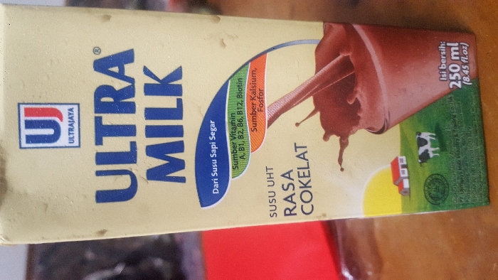 A beige-colored Ultra Milk carton, 250 ml (8.45 fl.oz.) in volume, stands upright, prominently centered. The carton displays the "ULTRA MILK" brand in large blue capital letters, with the "UJ" logo and "ULTRAJAYA" positioned above. The product is identified as "SUSU UHT RASA COKELAT" (UHT Milk Chocolate Flavor), made "Dari Susu Sapi Segar" (From Fresh Cow's Milk). Nutritional information highlights "Sumber Vitamin A, B1, B2, B6, B12, Biotin" and "Sumber Kalsium, Fosfor." Graphics on the label include a depiction of chocolate milk splashing into a glass, a rural scene with a single black and white cow, a small house with a red roof, and a yellow sun against a green landscape. A circular Halal certification logo is present. Micro-text at the base of the carton identifies the manufacturer as PT. Ultrajaya Milk Industry & Trading Co. Tbk., with the production facility located in Cimenyan, Bandung, Indonesia. The carton's surface exhibits visible condensation droplets. It rests on a brown, textured surface, possibly wood. To the left, a blurred red object or surface is partially visible, while the background remains indistinct and dark. No people are present. No specific actions or interactions are occurring within the visible scene.