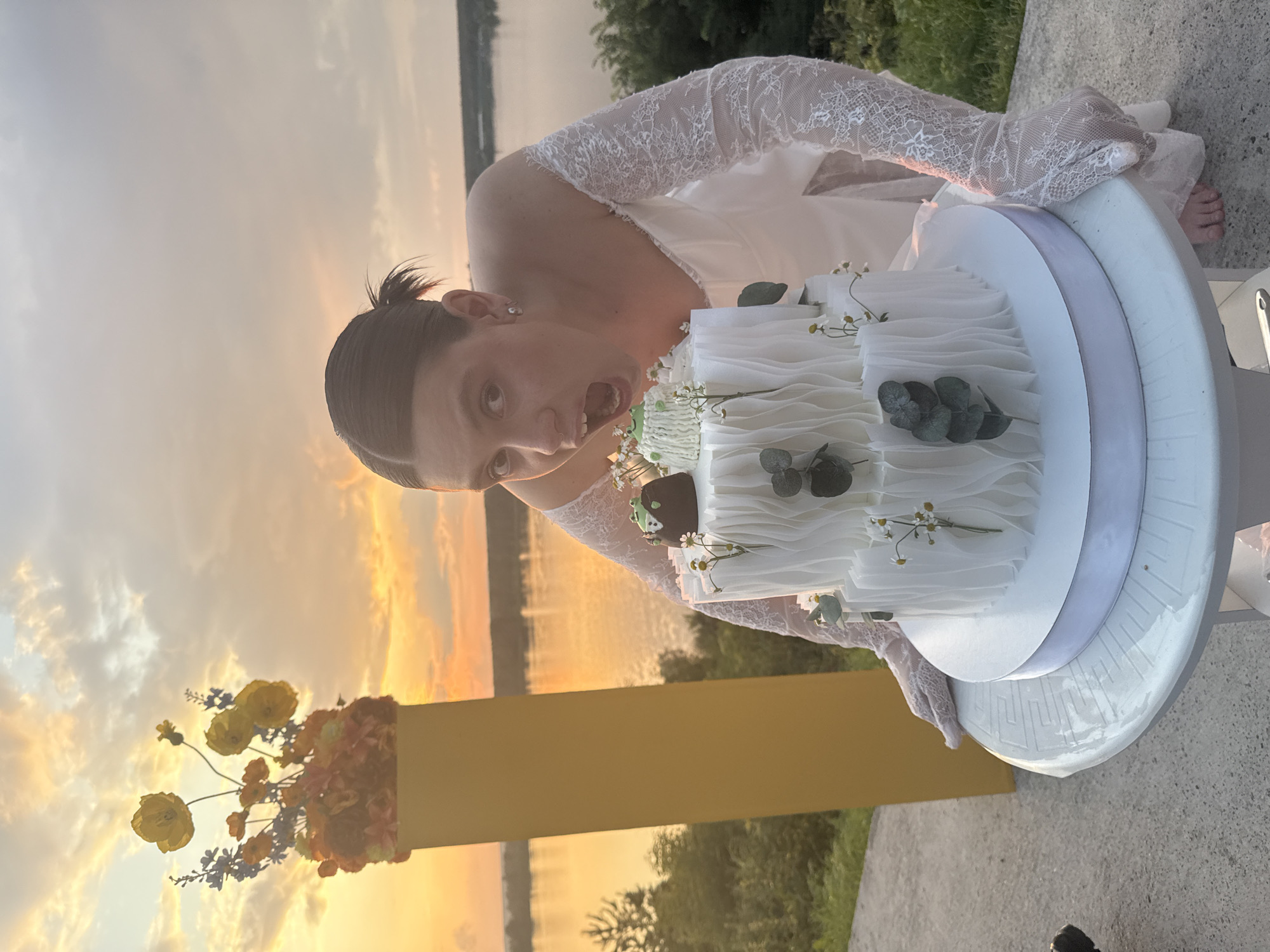 In Olkhovka, Russia, a young woman in a white off-the-shoulder dress and white lace gloves is captured mid-action as she leans towards a two-tiered white cake. The cake is adorned with small white flowers and eucalyptus leaves, with a decorative brown and green element on top. She is reaching out with her gloved hands to steady the cake, which sits on a white table. The setting appears to be outdoors during sunset, with a body of water and a tree line visible in the background. The sky is filled with warm, cloudy hues of orange and yellow. To the left of the frame, a tall yellow stand holds a bouquet of orange and yellow artificial flowers.