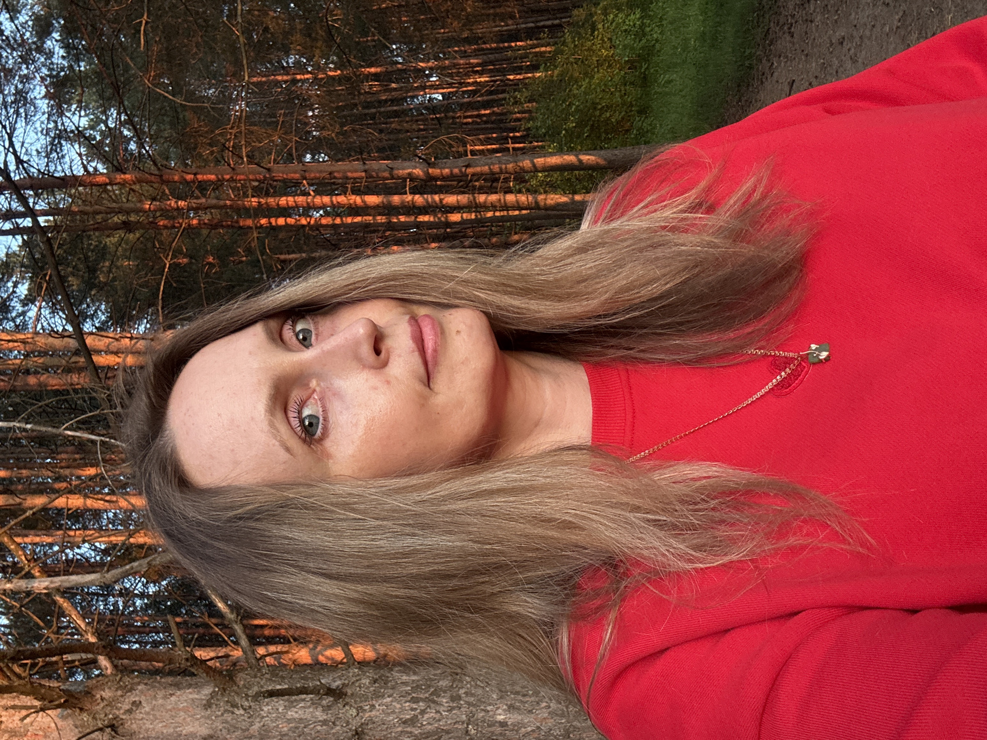 A woman with blonde hair and blue eyes is pictured outdoors in a forest. The sun is setting, casting an orange glow on the trees and the woman's face. She is wearing a red sweatshirt and a gold necklace with a green pendant. The background is a dense forest with tall pine trees. The ground appears to be a dirt path. The woman is looking towards the camera with a slight smile. The image was taken in the city of Chaikovsky, Russia.
