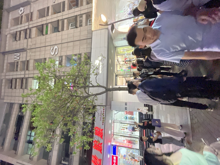 The image shows a busy city street in Hang Zhou Shi, China, located at coordinates 30.264 latitude and 120.156 longitude. A group of people are walking on the sidewalk in front of a large building. The building has a facade with the letters "W" and "S" prominently displayed. A large tree with green leaves grows in front of the building, and a street lamp casts a light over the scene. The building is adorned with windows, most of which have lights on inside, indicating that the scene takes place at night. The people are dressed in everyday clothing, and some are carrying shopping bags or other items. There are storefronts with signs in Chinese and English. Some of the storefronts are lit up with lights, and mannequins can be seen inside some of the windows. The people are walking in different directions, some looking at their phones. There is a sense of activity and energy in the air, indicating that this is a busy commercial district.