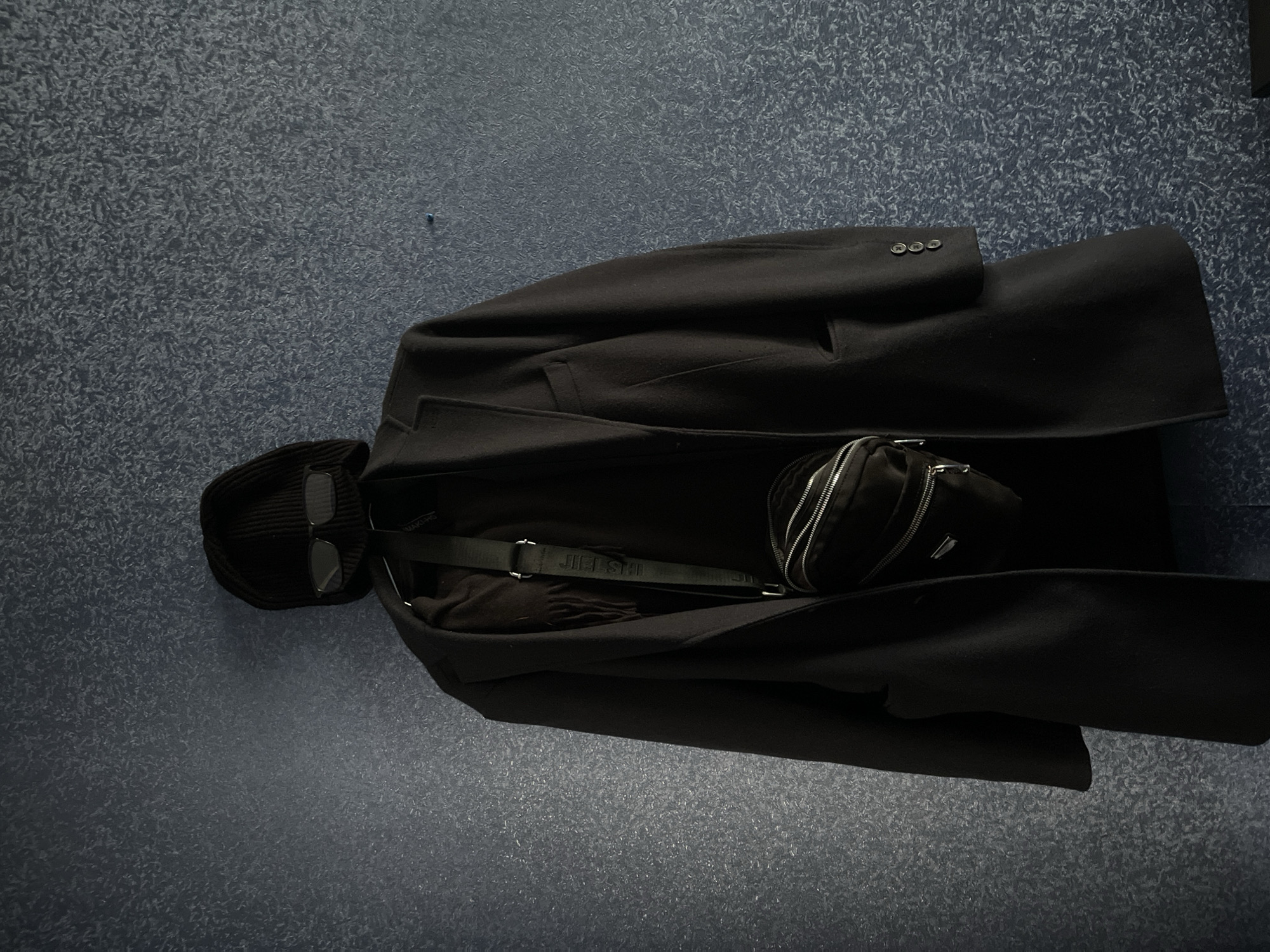 The image shows a black overcoat lying on a dark blue-grey surface with a speckled pattern. A black beanie hat with sunglasses on top of it is positioned to the left of the coat. A small black bag with a strap is partially visible on top of the coat. The strap has a visible brand name, "JIL.SHI". The location is not identifiable as Tbilisi, Georgia, based on the provided image alone.