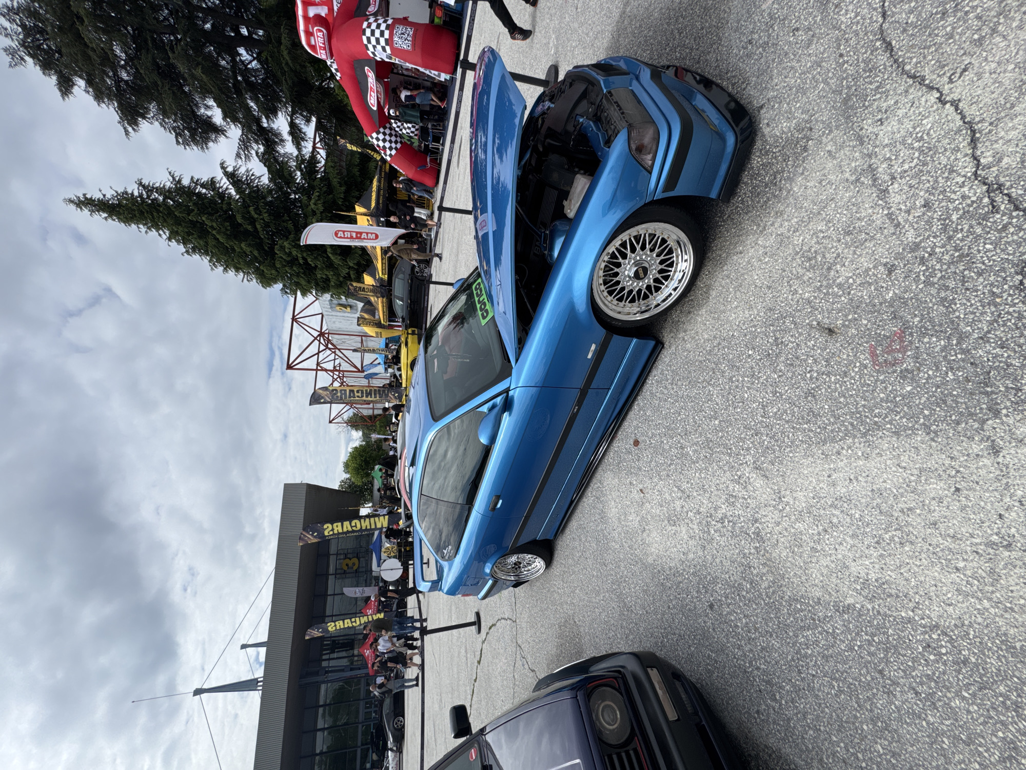 The image shows a blue BMW with its hood open at an outdoor car show. The car has custom wheels and a lowered suspension. Other cars and people are visible in the background, along with vendor tents and a building with signage. A tall evergreen tree and a cloudy sky are also visible. The location is not Plovdiv, Bulgaria.