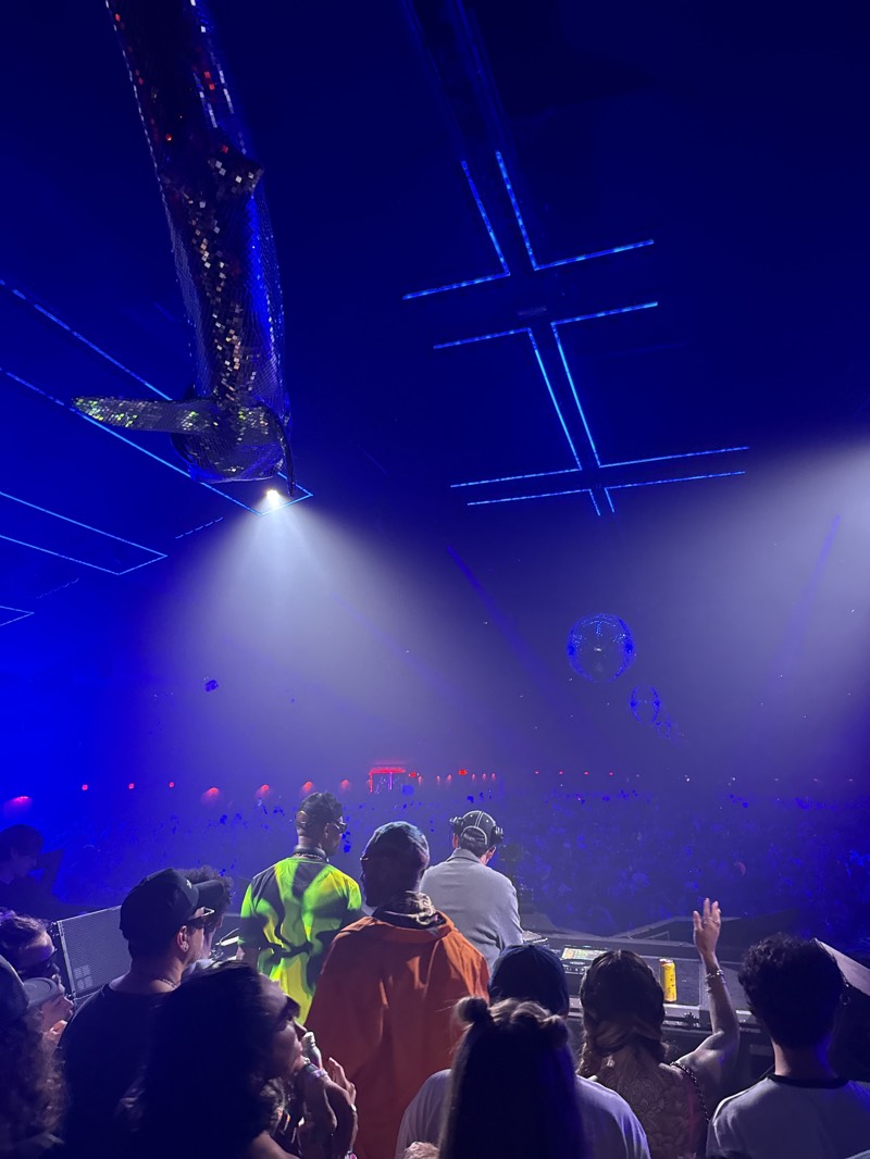 This image depicts a lively nighttime music event, likely a concert or DJ performance, held indoors within a large, dark venue. The perspective is from within the audience, looking towards a raised stage where performers are active.
In the foreground, a diverse crowd of people is visible, mostly from behind, facing the stage. Several individuals are distinct, including one wearing an orange hoodie and another in a vibrant neon yellow and black patterned shirt. These individuals are positioned close to the stage, observing the performers. Some attendees in the immediate foreground are holding drinks.
On the stage, at least two figures are present, operating what appears to be DJ equipment, including a mixing console or turntables. One performer wears headphones. The vast crowd extends into the background, illuminated by various stage lights, creating a sense of a large gathering.
The venue's ceiling is a prominent feature, adorned with intricate lighting and decorative elements. A large, elongated, sequined or mirrored object, possibly an artistic sculpture or illuminated airship, hangs suspended on the left, catching the light from a powerful spotlight. Elsewhere on the dark ceiling, multiple blue, glowing, cross-shaped light fixtures are arranged symmetrically. Several small, glittering disco balls are also visible, reflecting the ambient light.
The atmosphere is vibrant and energetic, characterized by strong blue and white spotlights, laser beams, and a general haze that diffuses the light, enhancing the immersive feel. The raised hands of some audience members suggest active engagement and enjoyment of the music. The indoor setting, artificial lighting, and large crowd are consistent with a music festival or club environment, aligning with the provided location context of Indio, United States. There is no visible text in the image.