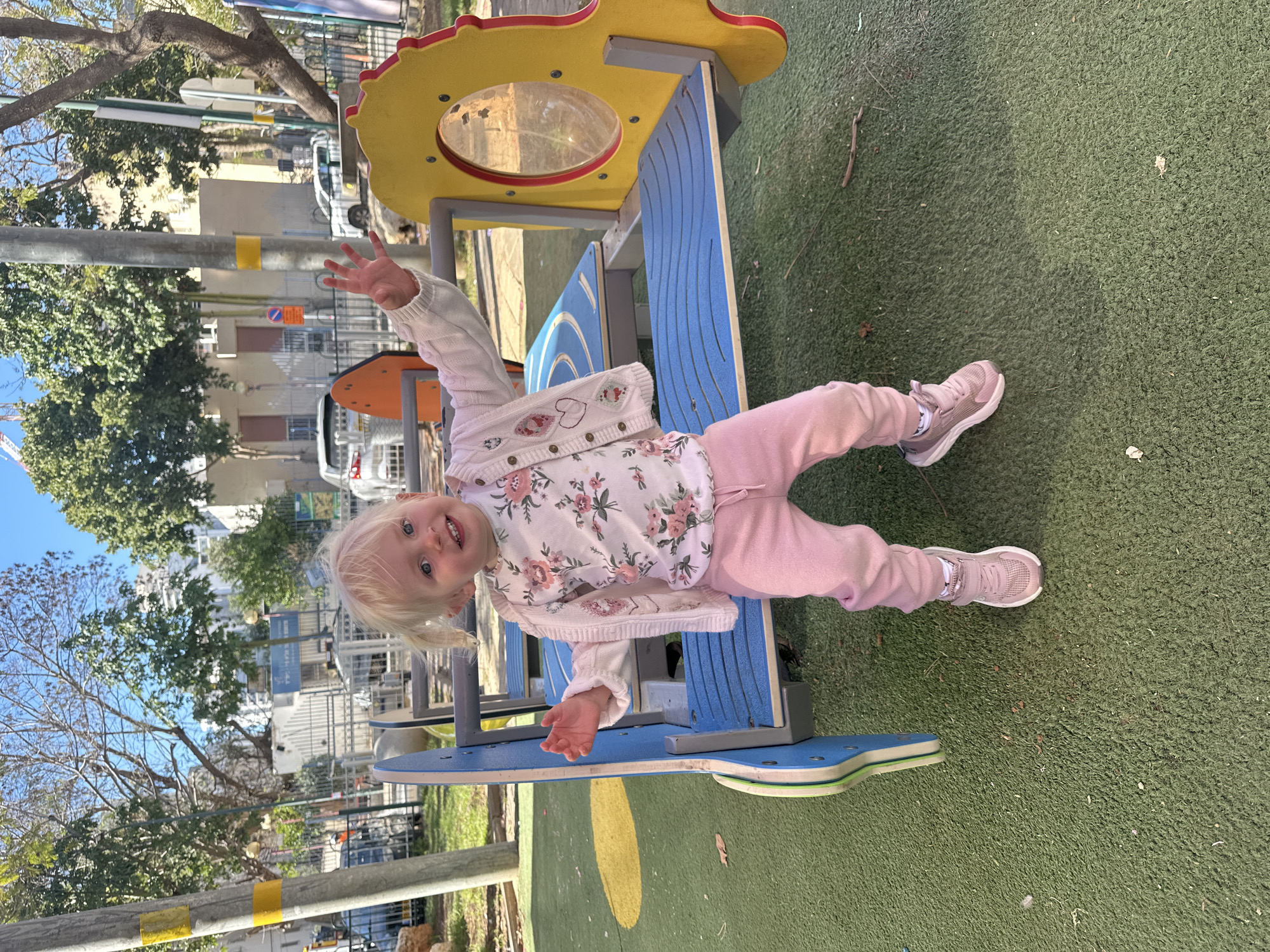 A young, blonde-haired child with blue eyes, appearing to be a toddler, is joyfully playing on an outdoor playground. The child is positioned on a vibrant blue and yellow piece of playground equipment, possibly part of a larger structure, which features a clear circular window. With a wide smile, the child looks directly at the camera and waves with both hands.
The child is dressed in a light pink floral long-sleeved top, a light pink knitted cardigan, light pink trousers, and matching light pink athletic shoes with white socks. The ground surface of the playground is artificial green turf.
In the background, under a bright, clear blue sky, there are trees (a mix of bare-branched and evergreen), residential buildings, and street infrastructure. A white car is visible in the distance, along with street poles. One pole features a yellow band, and another displays a red circular "No Parking" sign with Hebrew text underneath, indicating an urban or suburban setting likely in Israel.
The scene takes place during bright daylight on a sunny day, creating a cheerful atmosphere. The child's happy expression and playful gesture are prominent.