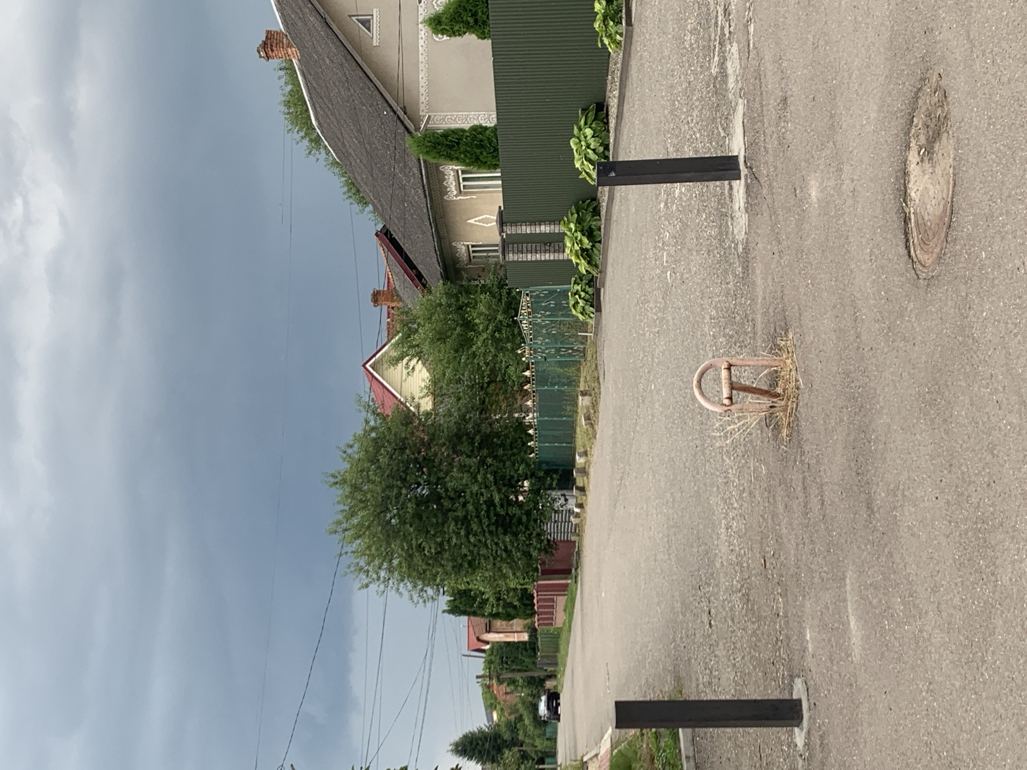 This is a vertical, eye-level, medium shot of a street in Kolomyia, Ukraine. The street is paved with asphalt and is lined with houses and fences. In the foreground, there are two black bollards on either side of the road, and in the middle of the road, there is a rusty metal object with dry grass growing around it. A black car is visible in the distance on the left side of the road, driving away from the camera. The sky is overcast and cloudy. The overall scene depicts a quiet residential street.