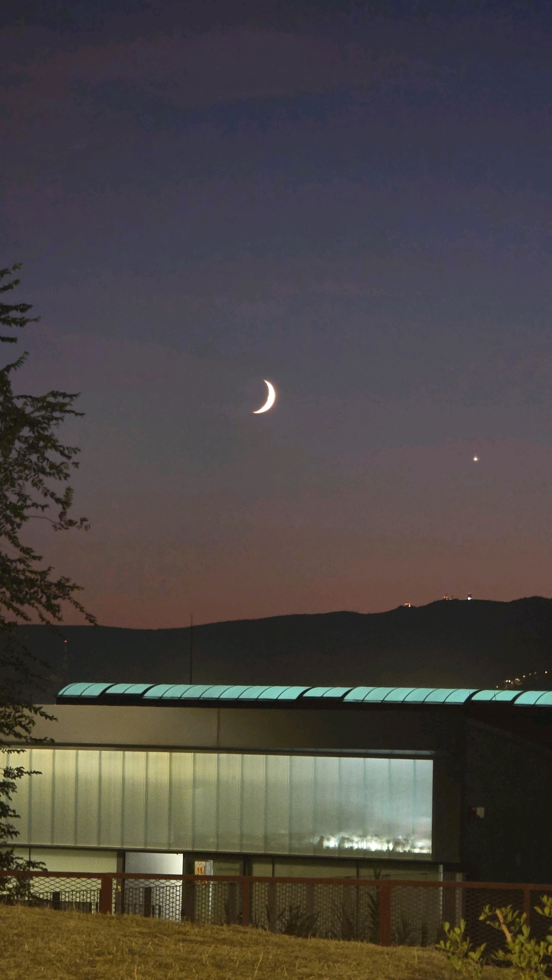 This is a photograph taken at dusk in Cornellà de Llobregat, Spain. The image captures a crescent moon and a bright planet in the twilight sky above a silhouetted mountain range. In the foreground, a modern building with a glass facade is visible, illuminated from within, with a dark railing in front of it. The ground in front of the building is covered in dry grass. A tree branch is partially visible on the left side of the frame. The overall impression is one of a peaceful evening in an urban or suburban landscape.
