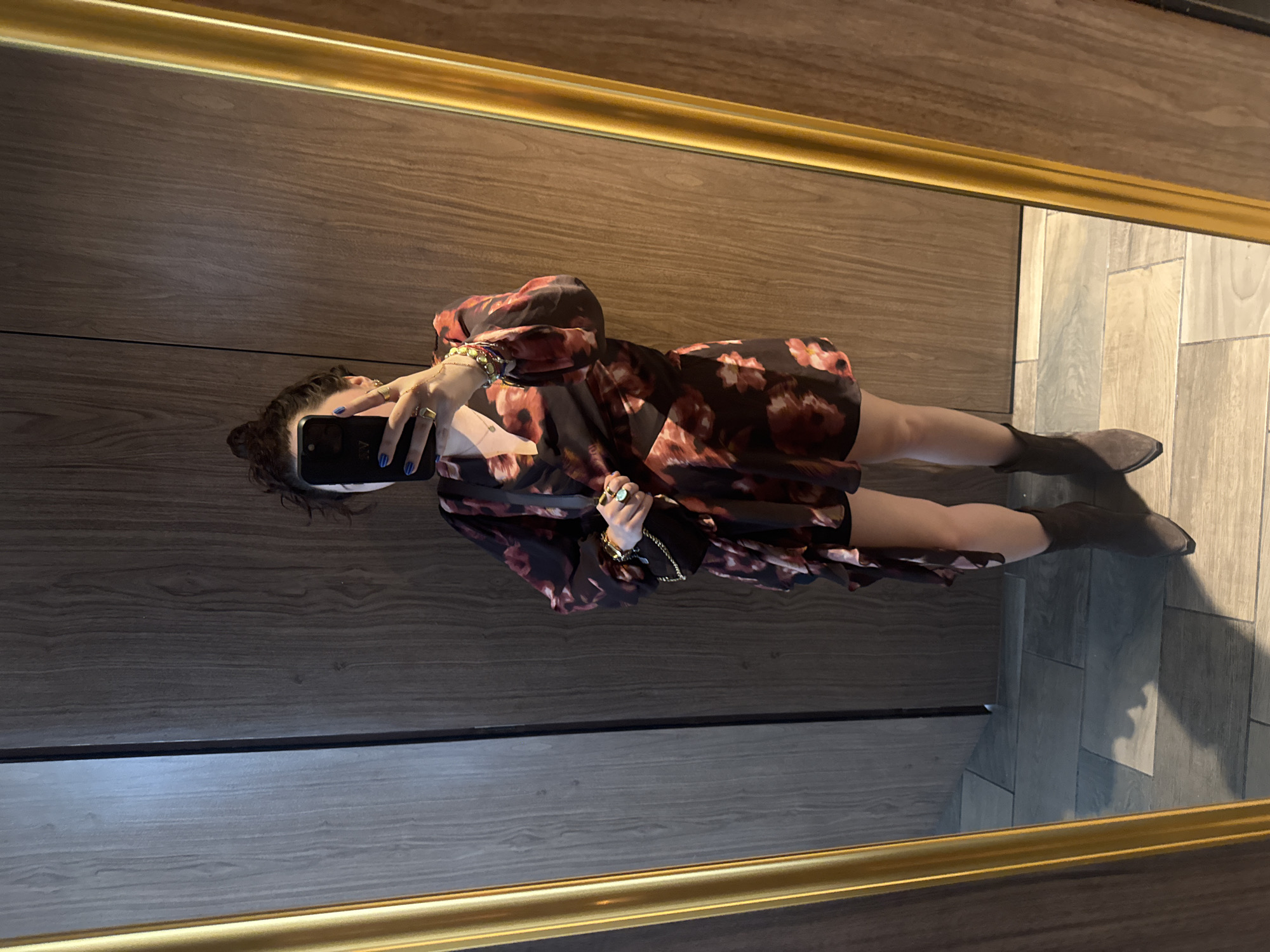 A woman stands in front of a mirror taking a selfie with a smartphone. The mirror has a gold frame. The wall behind the woman is a dark wood panel. She is wearing a short, floral-print dress and ankle boots. The floor is tiled. It's impossible to identify where this scene is located based on this image.