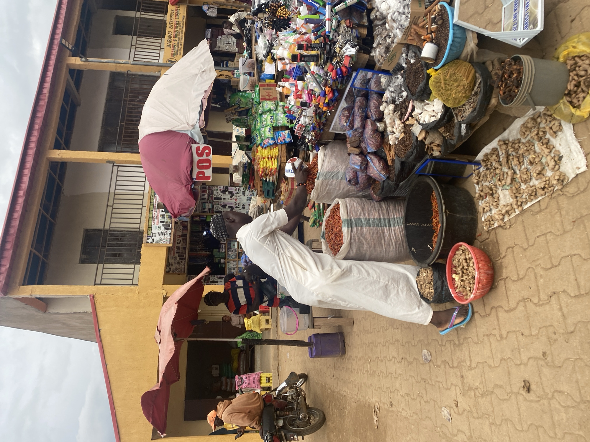 The image depicts an outdoor market scene in Kafanchan B, Nigeria, likely a bustling commercial area. In the foreground, a man dressed in a white robe is actively engaged in weighing dried goods from a large burlap sack into a white plastic container. Beside him, a red bowl overflows with dried chili peppers, and a black bucket also contains chili peppers. To his left, more dried goods are displayed, including what appears to be ginger spread out on a white cloth.
Behind him, a diverse array of items are on display, suggesting a general goods market. Shelves and tables are laden with packaged goods, bottles of cleaning supplies, toiletries, and various spices or dried foods. A sign reads "POS," indicating a point of sale system. Two large umbrellas, one red and one white and pink, provide shade over the stalls.
In the background, a person in an orange hoodie sits on a motorcycle, looking at a phone. Another person in a striped shirt is visible behind the man in white. The buildings in the background are multi-story structures with yellow and cream-colored facades and red roofing. The ground is paved with interlocking bricks. The overall atmosphere suggests a busy marketplace with a variety of goods and transactions taking place.