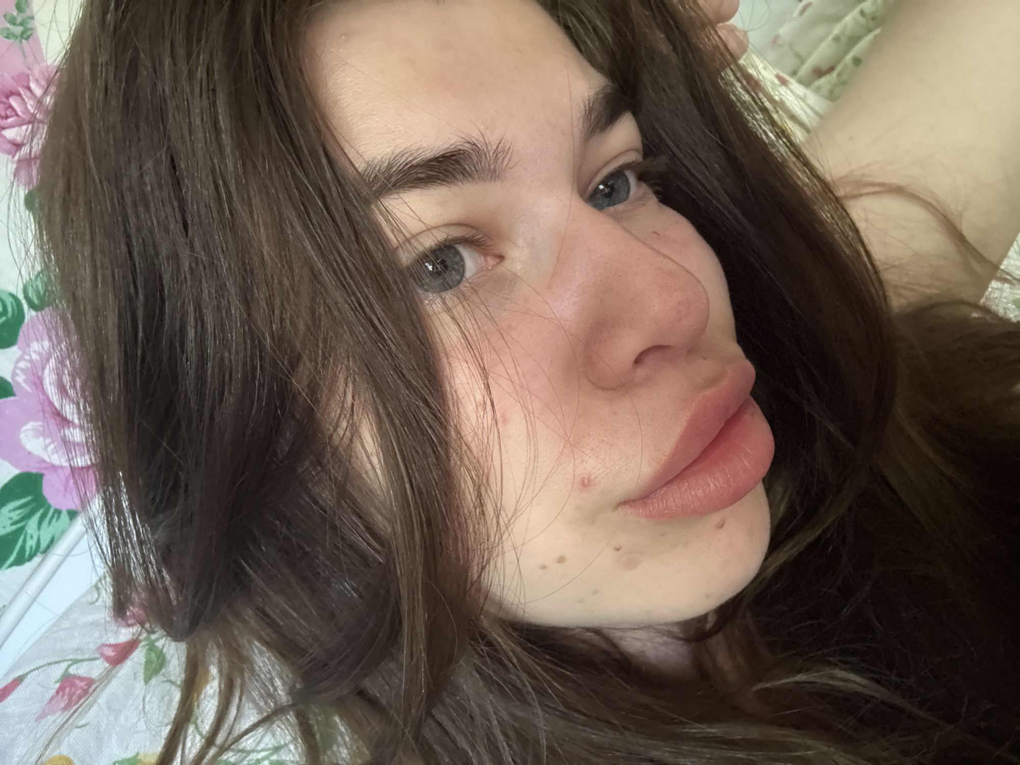 The image is a close-up, angled selfie of a young woman with long, dark brown hair that partially obscures her face. She has fair skin with some blemishes and freckles, and her eyes are a light grey-blue. Her lips are slightly parted and appear to be bare. The background is a floral-patterned fabric, likely bedding, with pink and green flowers. The lighting suggests it was taken indoors, possibly in a bedroom. The scene does not provide any information that would allow for the identification of a specific city or location within Мещёры, Russia.