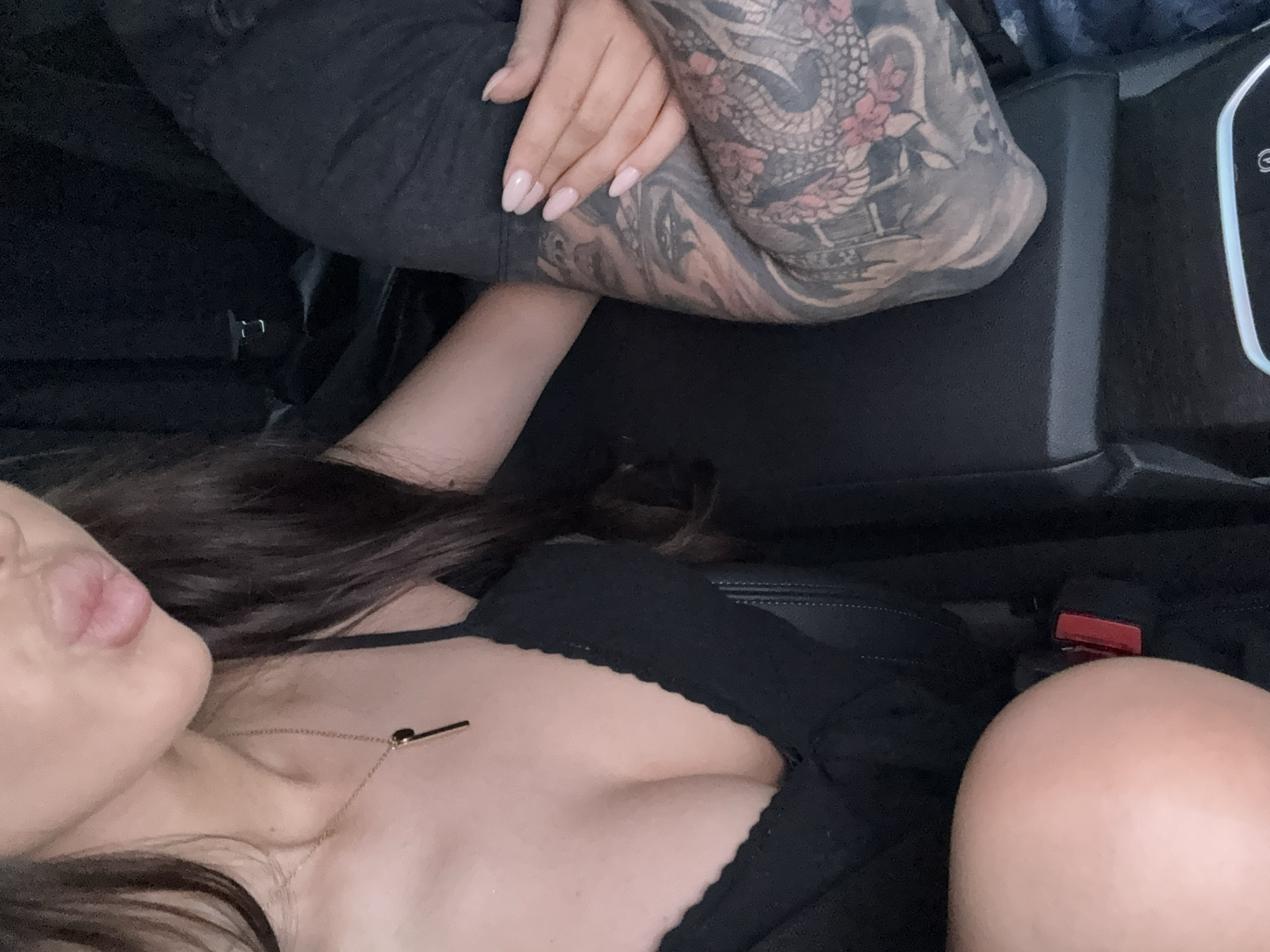 A close-up, tilted, high-angle shot shows a person in a vehicle. The person is wearing a black top with a scalloped edge, a delicate gold necklace, and is positioned in what appears to be a passenger seat. The seatbelt is fastened. Their arm, featuring a full-sleeve tattoo with designs resembling a dragon and flowers, rests on a black center console. The location is likely not in Manole, Bulgaria. The exact city is indeterminable without more information.