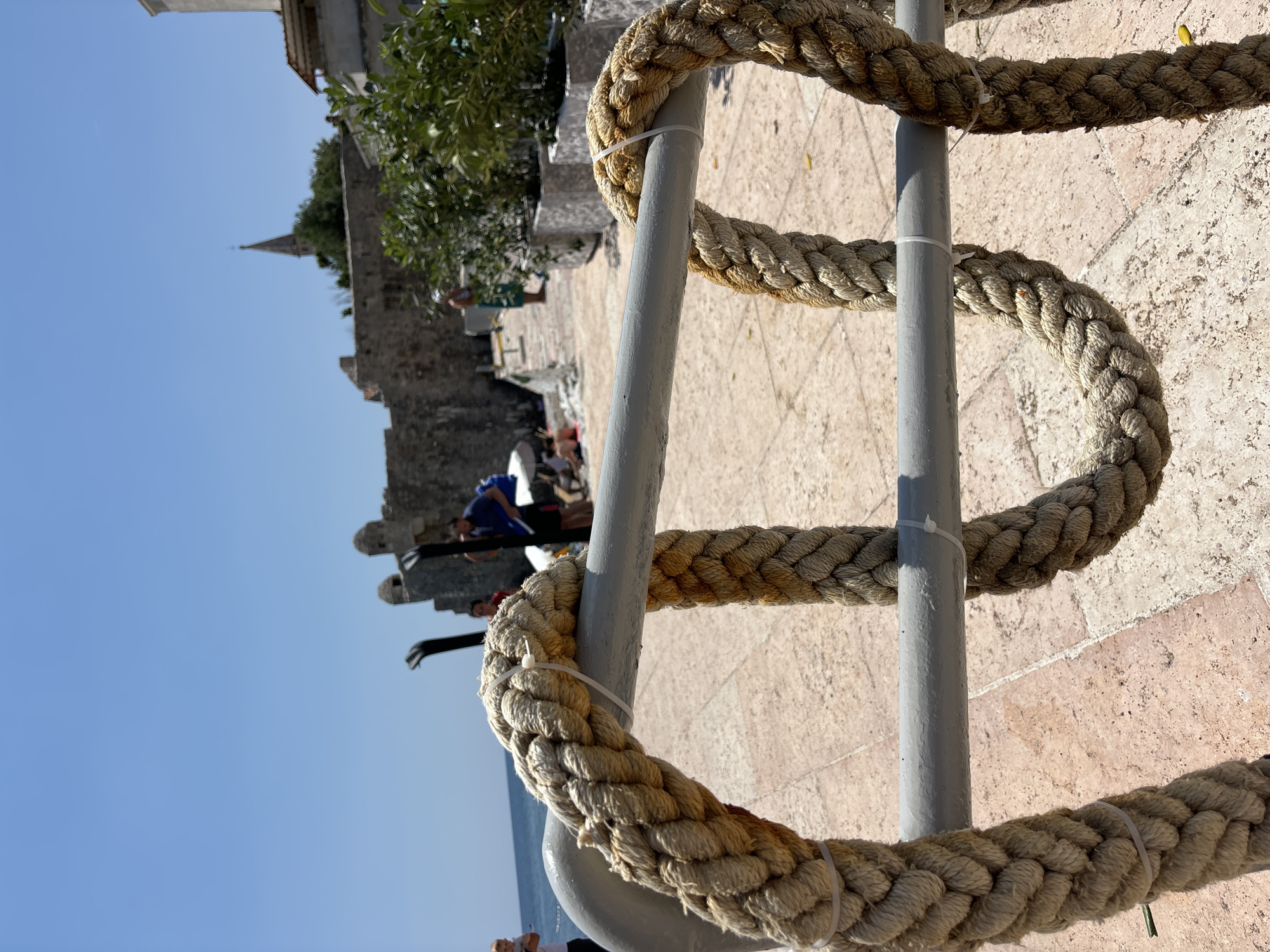 The image shows a close-up of thick, weathered rope coiled around a grey metal railing. The rope is secured with white zip ties. In the background, a stone wall and buildings, including a church spire, suggest a historical setting. The ground is paved with light-colored tiles. Beyond the wall, a clear blue sky and the sea are visible. A few people are casually scattered in the background, with one person wearing a blue shirt and another in red. The scene appears to be outdoors on a sunny day. The orientation of the image is rotated 90 degrees counter-clockwise. The overall impression is of a sunny day in a coastal town with ancient architecture.