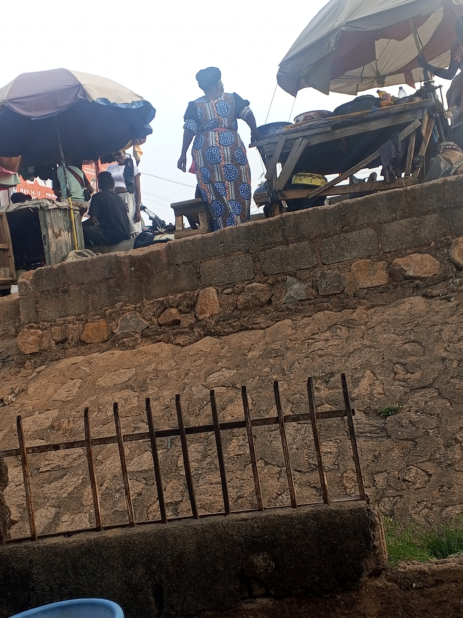 A photograph depicts a scene in Gwong, Nigeria. A woman in a patterned dress stands on a low wall, seemingly engaged with a vendor selling food from a wooden cart covered by a makeshift awning. The cart is laden with food items, obscured by the angle. Several people gather around the cart. A second awning-covered area indicates more vendors nearby. A rough stone wall, reinforced with a metal fence, dominates the foreground. The weather appears overcast.