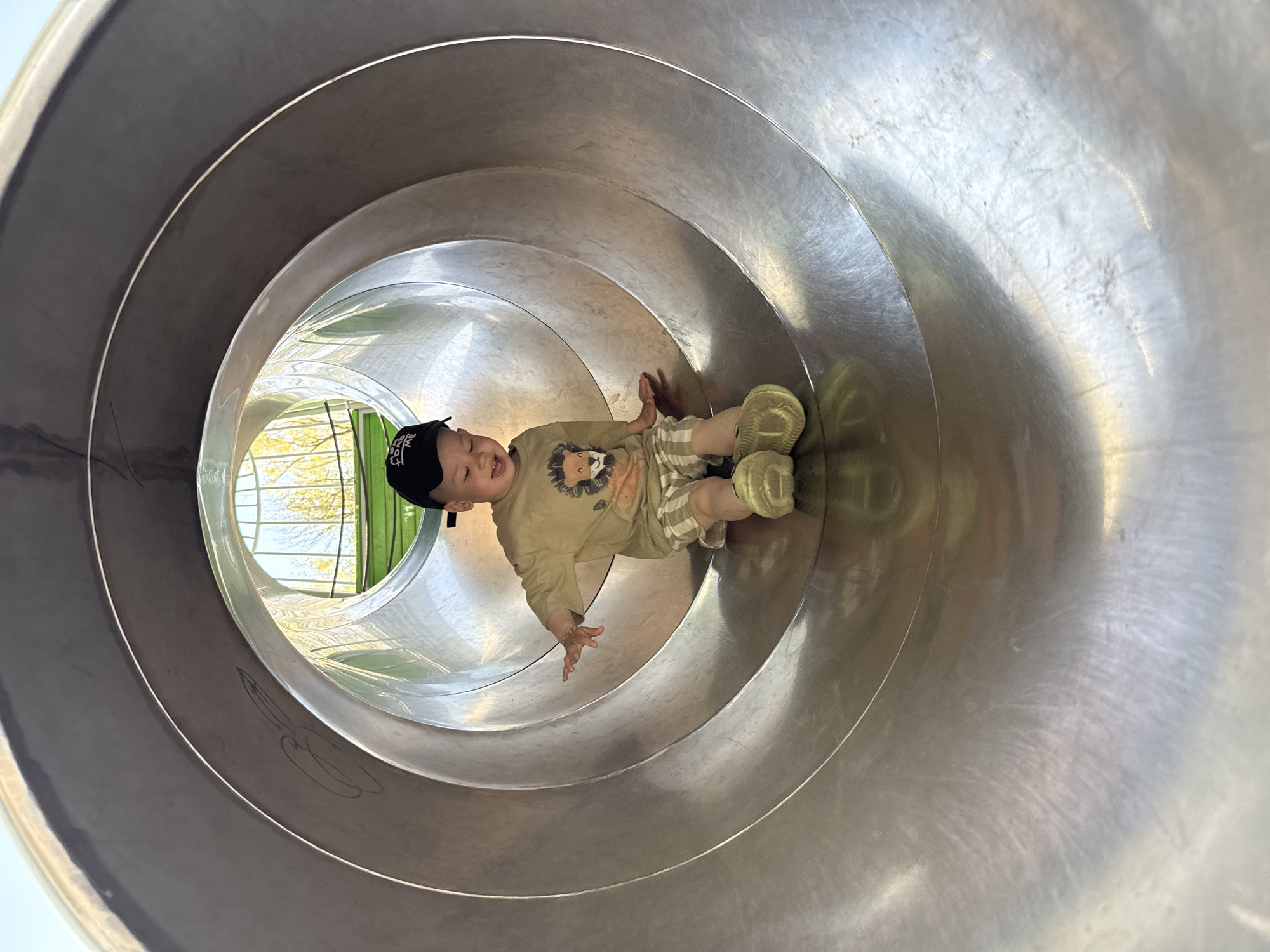 A toddler is inside a stainless steel tunnel slide, possibly at a playground. The child, wearing a black baseball cap, a tan shirt with a lion graphic, and checkered shorts with olive-green shoes, is sliding down the tunnel. The environment outside the slide appears green, suggesting trees or foliage. No specific details confirm the location as Rusanovo, Russia.
