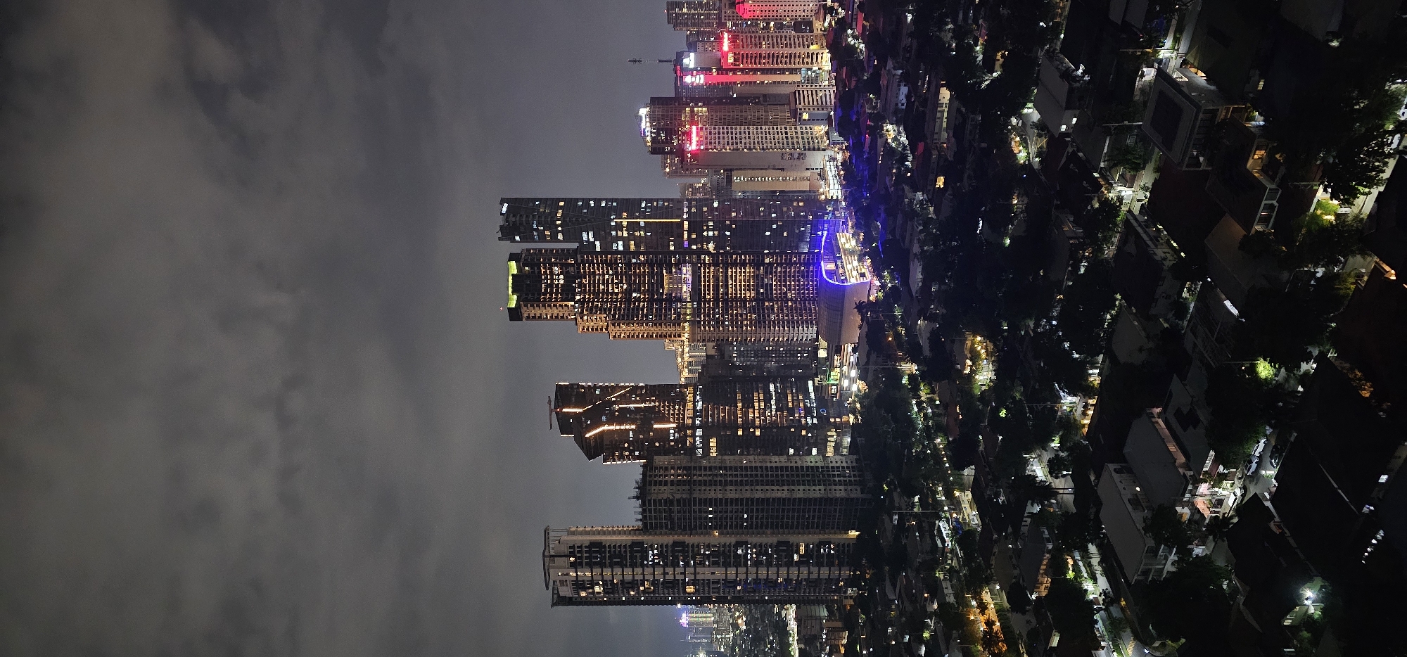 This is a nighttime, high-angle, vertical, full shot of the cityscape of Makati, Philippines. The image captures a dense urban environment characterized by numerous high-rise buildings illuminated with warm, yellow, and pink lights, creating a vibrant glow against the dark sky. The sky is overcast with heavy, gray clouds, obscuring any stars or celestial bodies.
In the foreground, a residential area with houses and trees is visible. The houses have distinct roof structures and are sparsely lit, with some windows showing interior lights. The trees are dark silhouettes, and streetlights cast pools of light on the roads.
The mid-ground and background are dominated by the modern architecture of Makati's business district. Several skyscrapers of varying heights and designs are prominently featured. Some of these buildings have intricate lighting patterns, with vertical and horizontal lines of light accentuating their facades. One building in the center appears to have a unique, curved architectural element illuminated with bright blue lights.
The overall impression is one of a bustling, developed metropolis at night, with a stark contrast between the bright, illuminated commercial structures and the darker, more subdued residential areas. The image appears to have been taken from a considerable height, likely from another tall building, offering a panoramic view of this part of Makati.