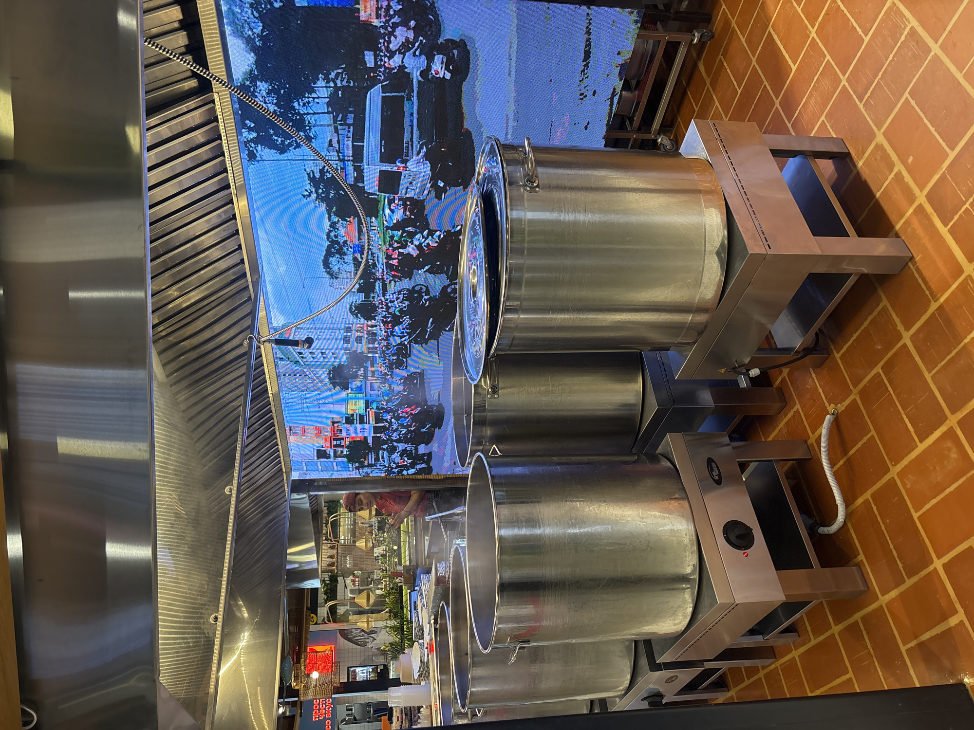 The photograph shows a commercial kitchen with stainless steel cooking equipment, including large stock pots on stands, positioned beneath an exhaust hood. A large video screen in the background displays scenes from what appears to be a street in Saint Petersburg, Russia, showing traffic and pedestrians. The kitchen floor is tiled in a reddish-brown pattern. A person wearing a red shirt and a bandana is visible working in the background of the kitchen.