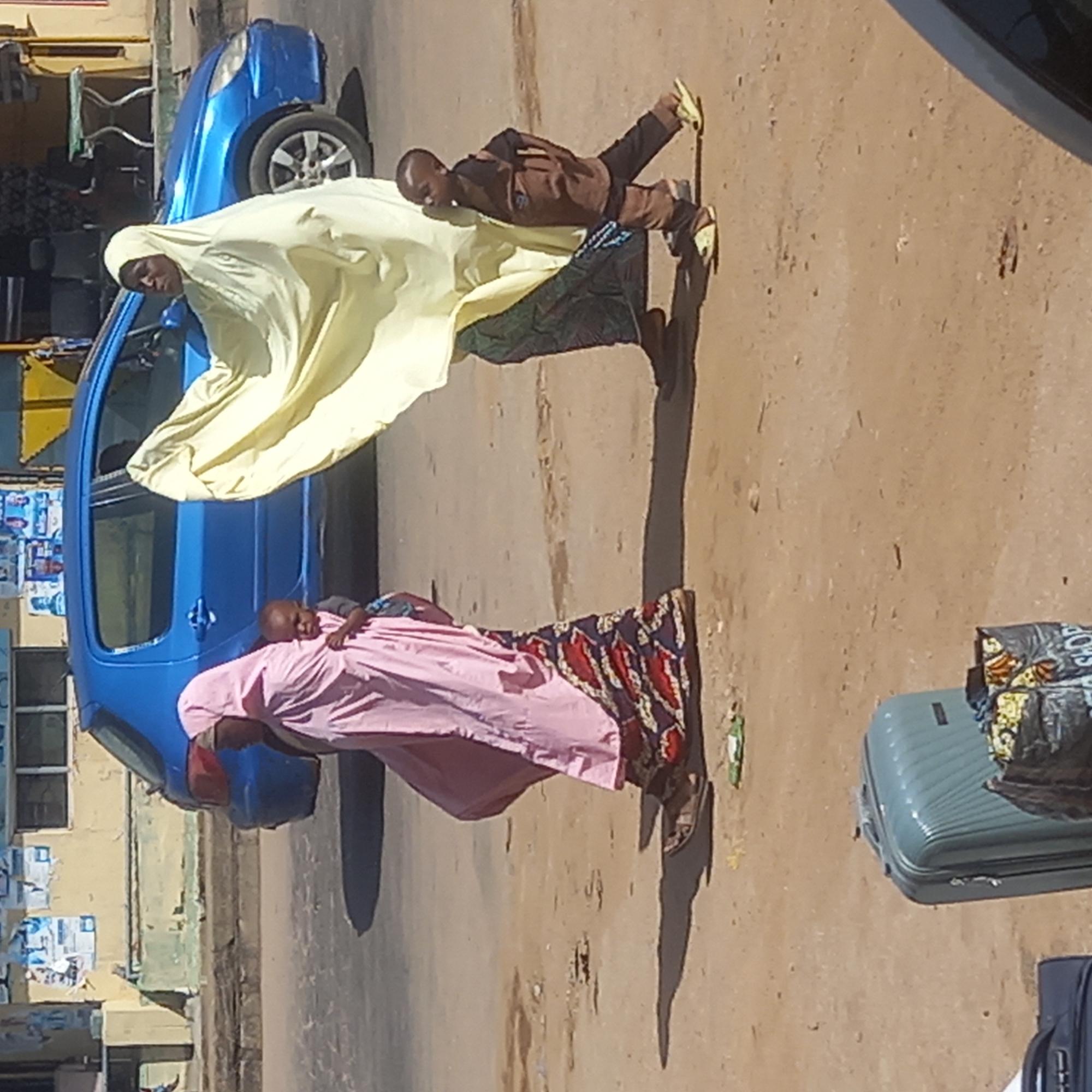 The image depicts a daytime street scene in Sardauna, Nigeria, featuring several individuals and a parked blue car on an unpaved road. The sunlight is bright, casting distinct shadows, suggesting it's either mid-morning or late afternoon.
In the foreground, on the left, a woman is seen standing near the blue hatchback car. She wears a light yellow, flowing head covering that drapes over her body. Her face is visible, and she looks towards the viewer. To her right, a young child stands, dressed in a brown long-sleeved top, dark trousers, and light yellow shoes. The child appears to be interacting with or standing very close to the woman in yellow.
Further to the right, another woman is present, distinguished by her pink head covering and a pink garment worn over a patterned skirt or dress. She is carrying a baby on her back, who is partially visible and wearing a striped top.
On the far right side of the frame, two pieces of luggage are visible on the ground. One is a light blue/grey hard-shell suitcase, and on top of it rests a black bag with a colorful pattern, where faint text like "SHOP" can be discerned. Another dark suitcase is partially visible below the light blue one.
In the background, behind the car, there is a building with light-colored walls and windows. Several posters or flyers are affixed to the building's facade, though their text is illegible. The ground throughout the scene appears to be dirt or sand, indicating an unpaved road or shoulder. The overall impression is one of people gathered or waiting by the roadside, possibly preparing for travel.