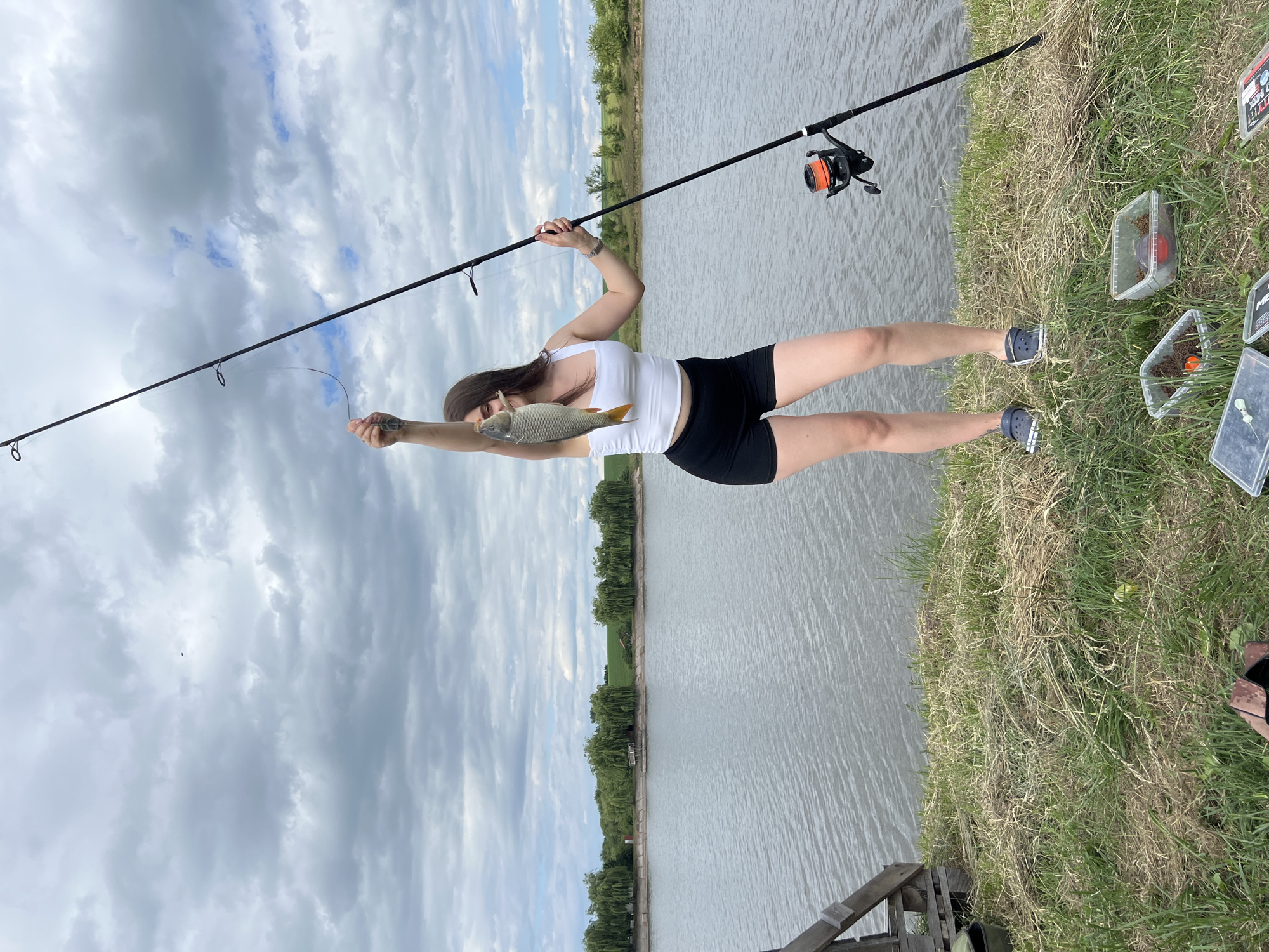 A woman is fishing on the bank of a body of water, likely a lake or river, with trees and greenery in the background under a cloudy sky. She is holding up a fish she has caught. A fishing rod is propped up beside her. The scene takes place on the outskirts of Borychivka, Ukraine.