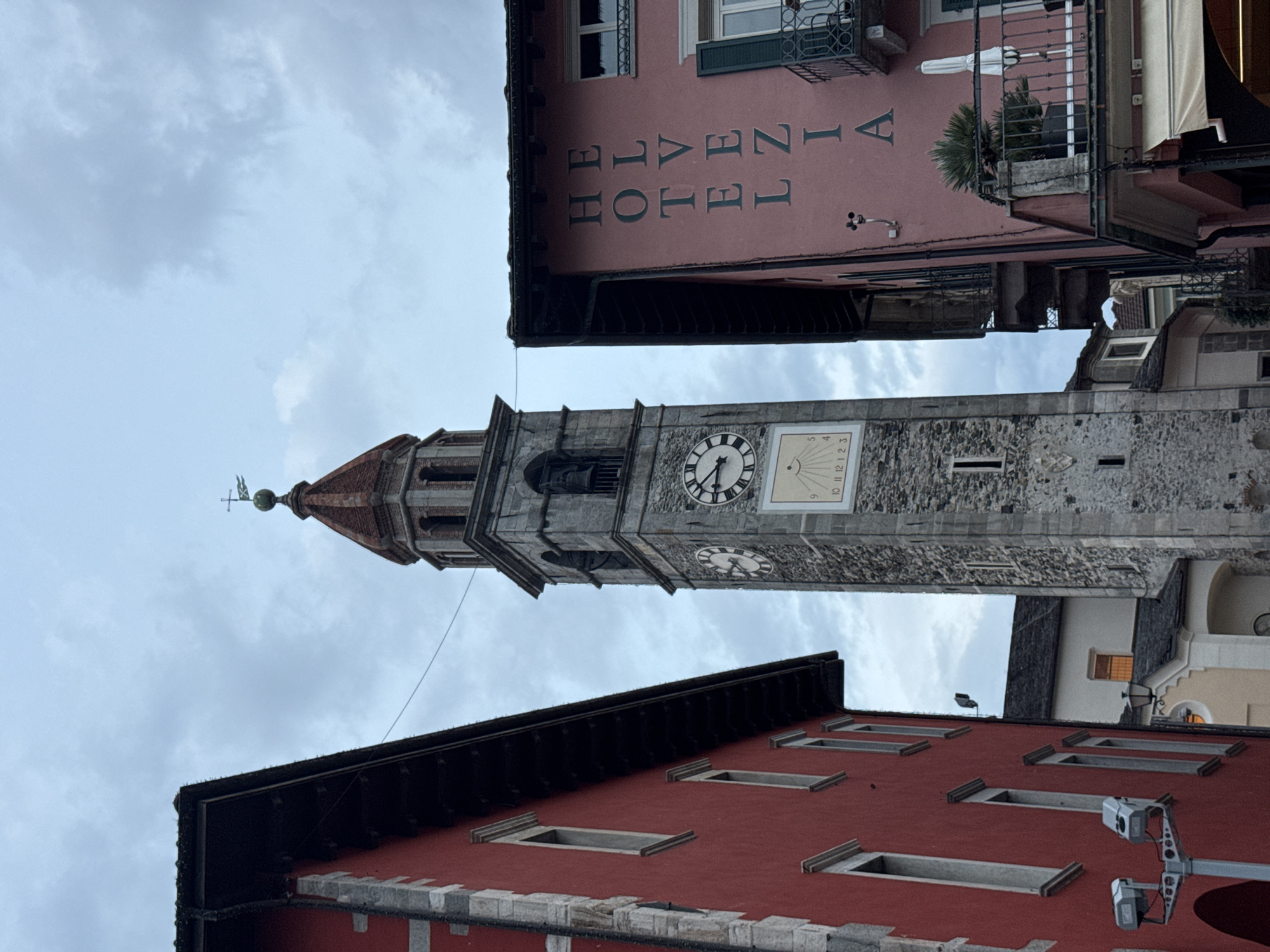This is a low-angle, full-shot photograph of a clock tower and two buildings in the town of Ascona, Switzerland. The clock tower, which is made of stone, is located in the center of the frame and has a red-tiled roof. There are two clocks on the tower, one on each side, and a sundial below them. The building on the left is painted red and has several windows. The building on the right is painted pink and has the word "HOTEL HELVEZIA" written vertically on its facade. There are balconies and awnings on the pink building. The sky is overcast. The image appears to have been taken during the daytime. There are no people visible in the image.
