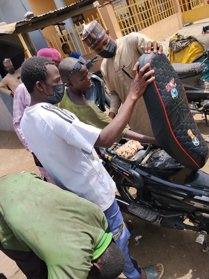 The scene is set outdoors in Fagge D2, Nigeria, under bright daylight. Five individuals are gathered around a black motorcycle. A man in a light brown long-sleeved garment, patterned cap, and black face mask holds the rear of a black motorcycle seat, which features a hexagonal pattern, red trim, and colorful logos. A second man, wearing a white t-shirt with black stripes on the sleeves and a black face mask, holds the front of the same detached seat. A young boy in an olive green shirt with sunglasses perched on his head stands between them, observing the motorcycle. The motorcycle's under-seat compartment is exposed, revealing internal components and a crumpled light brown fabric. In the lower-left foreground, a third man in an olive green shirt is bent over, facing downwards. In the background, a man in a light yellow shirt with a patterned cap and mask stands with hands on his hips, observing. Light-colored buildings with metal grates and other partially visible motorcycles are present in the background.