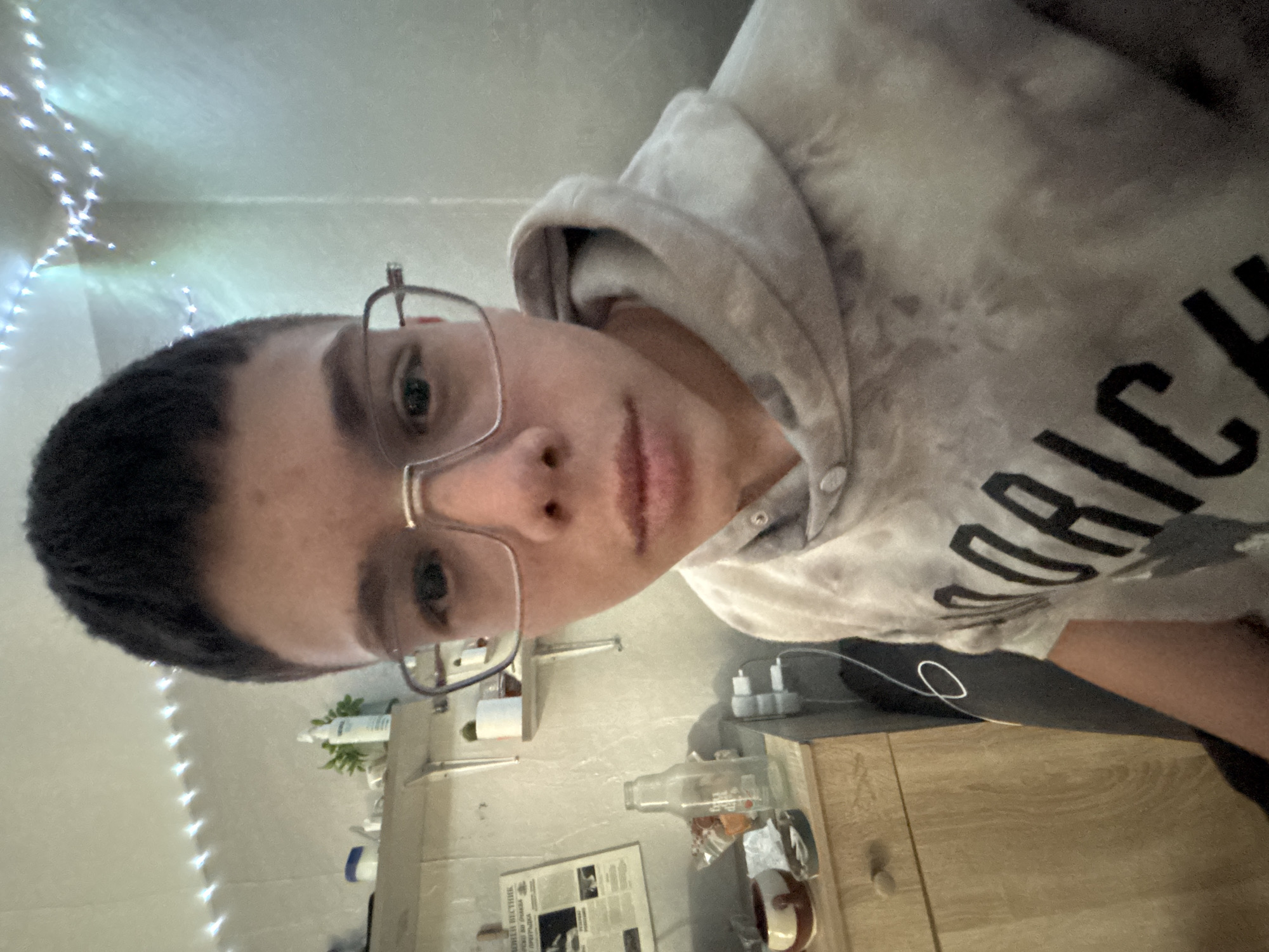 A close-up, angled selfie shows a person with short, dark hair, fair skin, and wearing eyeglasses. They are dressed in a light-colored hoodie with dark lettering visible. The background appears to be a room with off-white walls adorned with string lights. A countertop or shelf is visible with various objects including bottles, toiletries, a newspaper, and a mug. Electrical outlets and cords are also present. The image location information (Troyan, Bulgaria) does not appear related to the image.